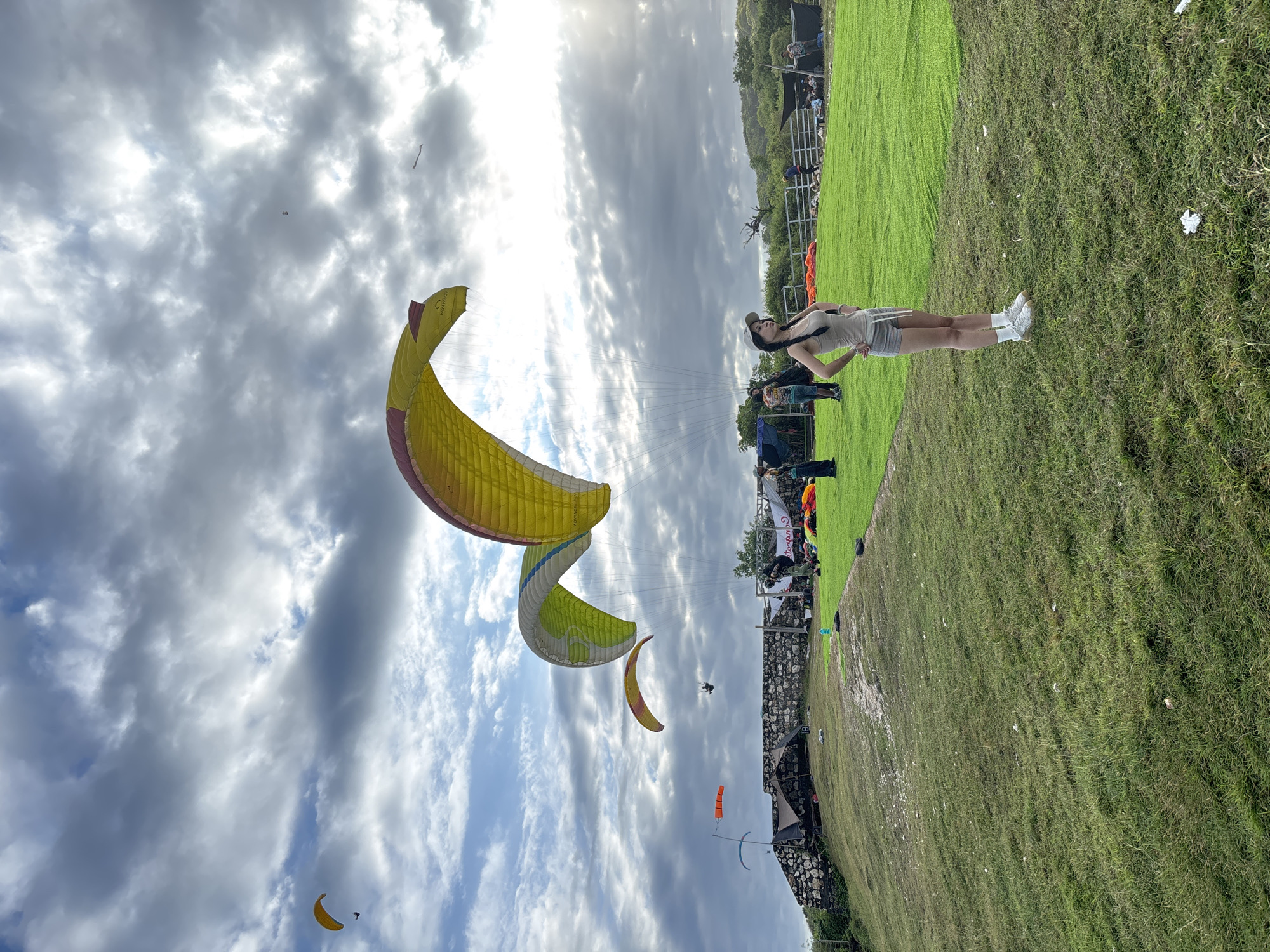 The image depicts a scene at Timbis Paragliding in Kuta Selatan, Indonesia. In the foreground, a woman stands on a grassy field with her hands on her hips, watching paragliders in the air. The sky is cloudy with patches of blue, and the sun is partially obscured. Several colorful paragliders are visible against the sky. A group of people, a fence, flags, and tents are visible in the background.