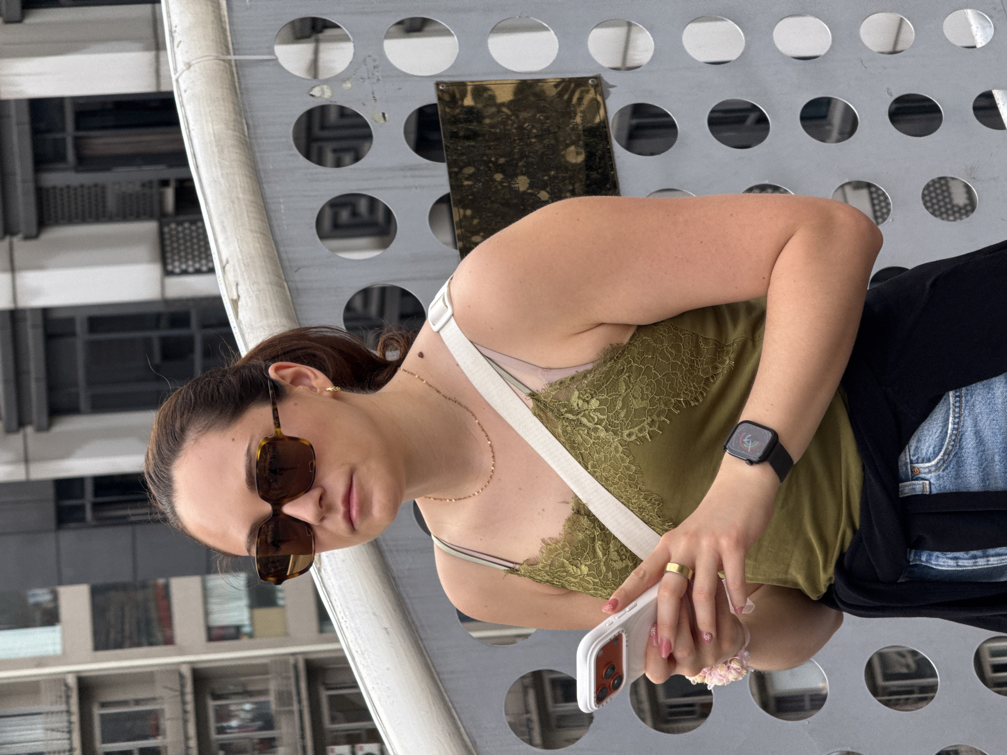 A woman is standing in Yuzhong District, China, holding a cell phone. She is wearing sunglasses, an olive green lace-trimmed tank top with a white strap, a gold necklace, a black sweater, and denim shorts. She also has a black smart watch on her left wrist. The background features a metal structure with circular cutouts and a building with window frames.