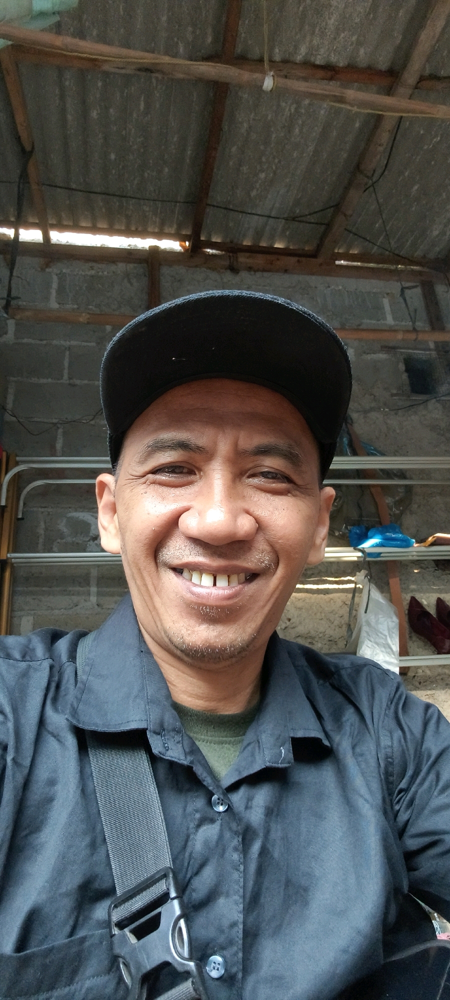 In South Tangerang, Indonesia, a middle-aged man with a dark baseball cap and a dark collared shirt is seen in a close-up, smiling selfie. The man has a light complexion, short dark hair, and a goatee. He is wearing a dark collared shirt over a green t-shirt, with a dark strap across his chest. The background appears to be a workshop or storage area, with a corrugated metal roof supported by wooden beams. Gray cinder block walls are visible behind him, and shelves holding various items, including what appear to be shoes, are present. The lighting suggests it is daytime.