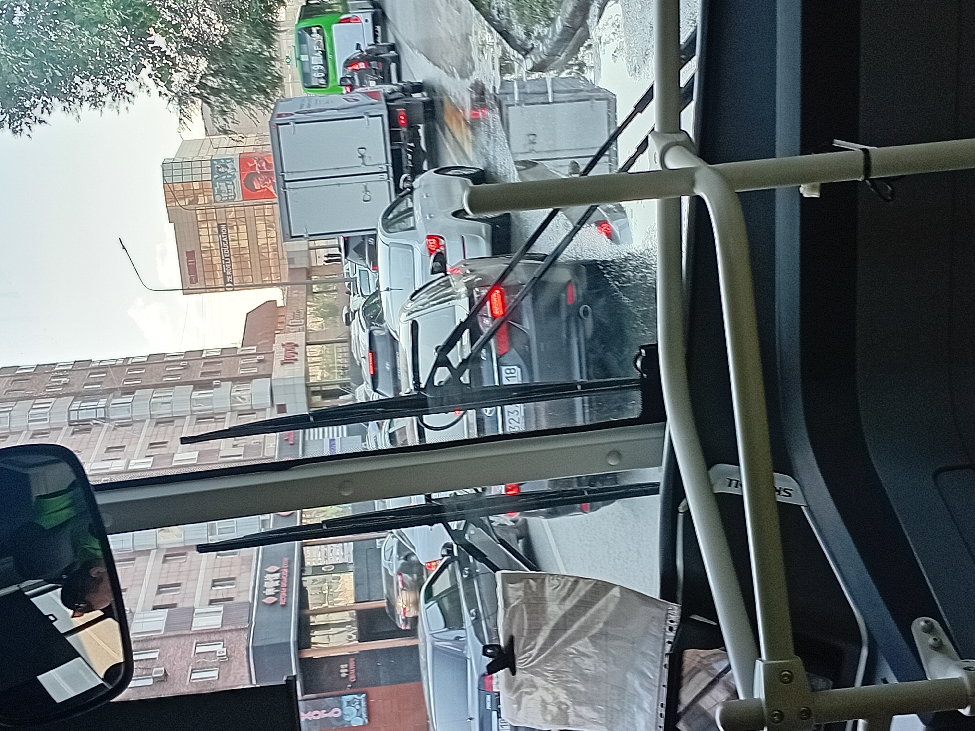 The image shows a traffic jam in the city of Semey, Kazakhstan. In the foreground, we see the interior of a vehicle, possibly a bus, with a view through the windshield. Two windshield wipers are visible. Outside, several cars are lined up on a wet road, reflecting the overcast sky and surrounding buildings.
One of the cars in the traffic jam has a license plate with the numbers "323 18". A green bus with a license plate "6 91A F 16" is also visible in the distance, alongside a white truck. The buildings in the background appear to be apartment blocks and commercial establishments, with some signs in Cyrillic script. The weather appears to be overcast and possibly raining, as evidenced by the wet road and reflections.
In the rearview mirror of the vehicle in the foreground, a person wearing sunglasses and a green vest is visible, presumably the driver. The overall scene depicts a typical urban traffic situation in Semey, Kazakhstan, with cars and buses navigating congested streets.
