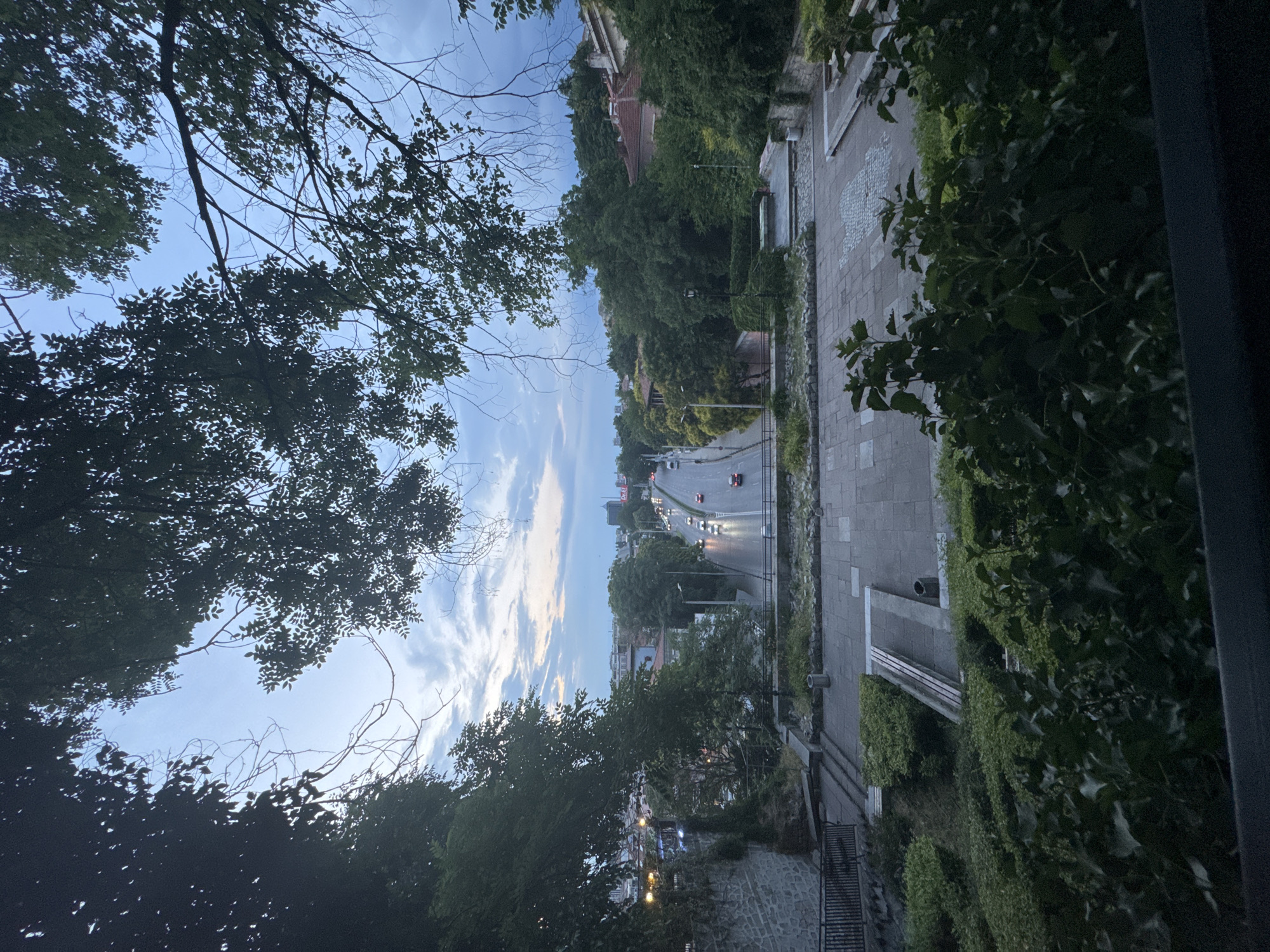 This image depicts an outdoor scene in Plovdiv, Bulgaria, likely during the late afternoon or early evening. The perspective is from an elevated position, looking down towards a street lined with trees and buildings. In the foreground, large trees with dark green foliage frame the view, with their branches silhouetted against a partly cloudy sky. The sky itself transitions from a pale blue near the horizon to a darker shade above, with streaks of white and light orange clouds suggesting the setting sun.
The street below shows moderate traffic, with several cars visible. The road appears to be a main thoroughfare, with buildings and dense greenery on either side. A paved walkway or sidewalk runs alongside the street, featuring a stone-like pattern. Further down the street, there are hints of urban development with taller buildings.
The overall atmosphere is calm and serene, with the natural elements of the trees and sky contrasting with the man-made street and buildings. The lighting suggests a transitional time of day, possibly before dusk, casting a soft glow over the scene. There is no visible text in the image.
