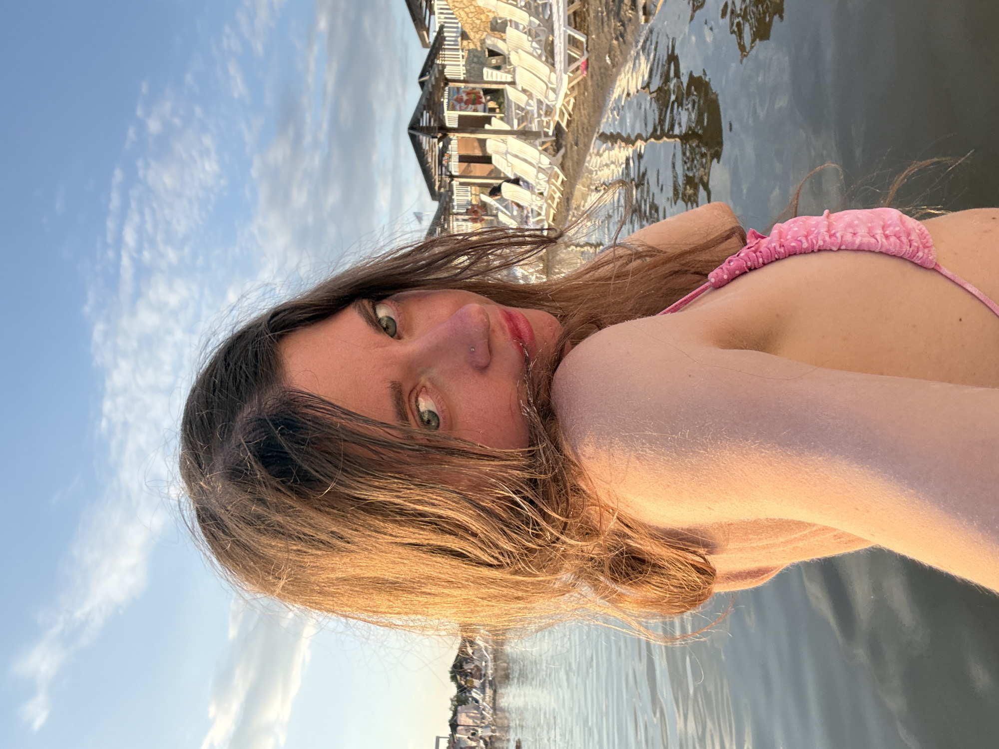 A close-up, vertically oriented selfie shows a young woman with light brown hair, some of which falls over her face. She is wearing a pink bikini top with white polka dots. Her green eyes are visible, and she has a nose ring. The woman appears to be in the water, with the reflection of buildings and a cloudy sky visible on the surface. In the background, there are beach chairs and awnings, suggesting a beach resort setting. The lighting indicates it is likely late afternoon or early evening. The image was taken in Sol-Iletsk, Russia.
