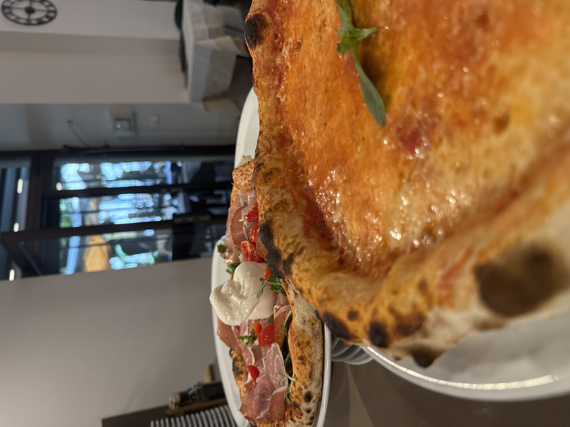 Two pizzas are presented on white plates, with the foreground pizza slightly out of focus and the background pizza in focus. The foreground pizza is a Margherita style pizza, with a red tomato sauce base and melted cheese, garnished with a single basil leaf. The background pizza is topped with prosciutto, cherry tomatoes, arugula, and a dollop of fresh mozzarella cheese. The pizzas are placed on a table in what appears to be a restaurant. In the background, through a glass door, a blurred interior of a restaurant with tables and chairs is visible. The scene is indoors. The location in the city of Trento, Italy is not explicitly identifiable from the image.