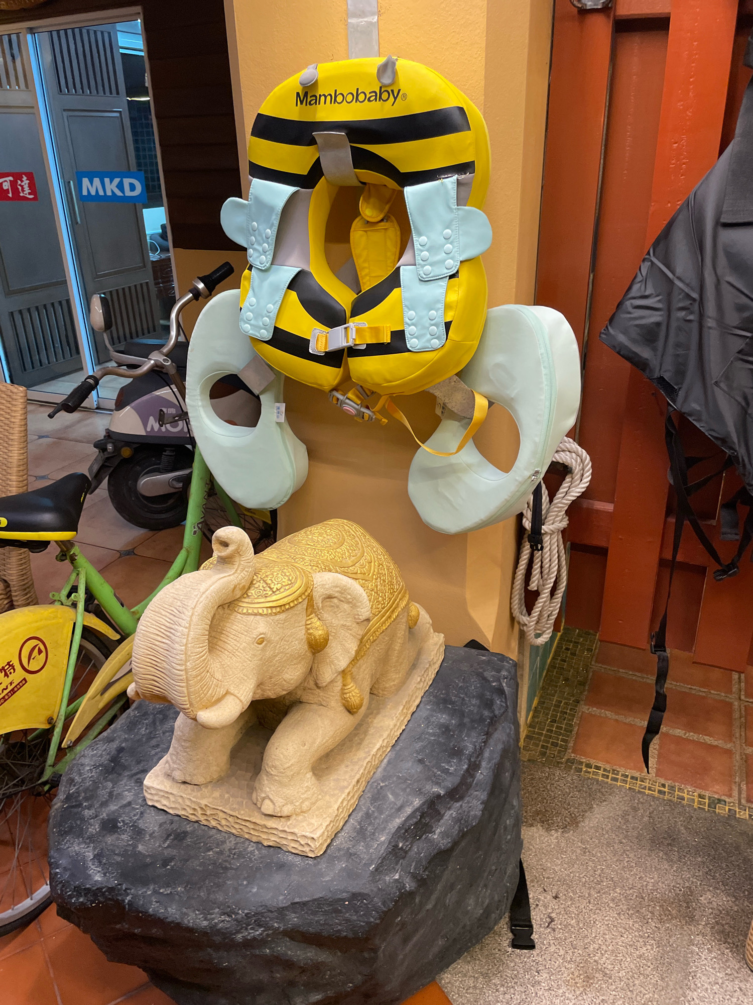 In Pattaya City, Thailand, an outdoor scene displays a variety of objects. A yellow and black inflatable swim ring, designed to resemble a bee and branded "Mambobaby," is prominently displayed. Attached to the swim ring is a light blue floatation device with snap closures. Beside the swim ring, a black jacket hangs from a support structure, and a thick rope is tied to the floatation device. In the foreground, a beige stone elephant statue is positioned on a large, dark grey rock. A green bicycle with a yellow basket and black seat is visible to the left of the elephant, partially obscuring a portion of the statue. The background features a doorway with glass panes and a sign that reads "MKD." The overall environment suggests a display area, possibly at a shop or market.