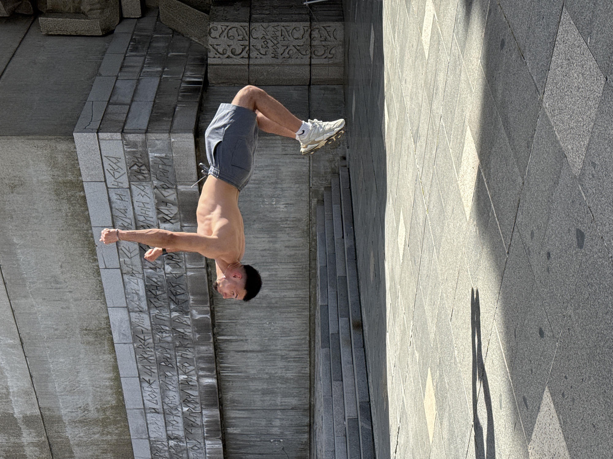 A man is performing a backflip in front of the Monument to 1300 Years of Bulgaria in Shumen, Bulgaria. The man is wearing grey shorts and white sneakers. His back is facing the viewer. The monument is made of grey stone and has inscriptions on it. The ground is paved with grey tiles.