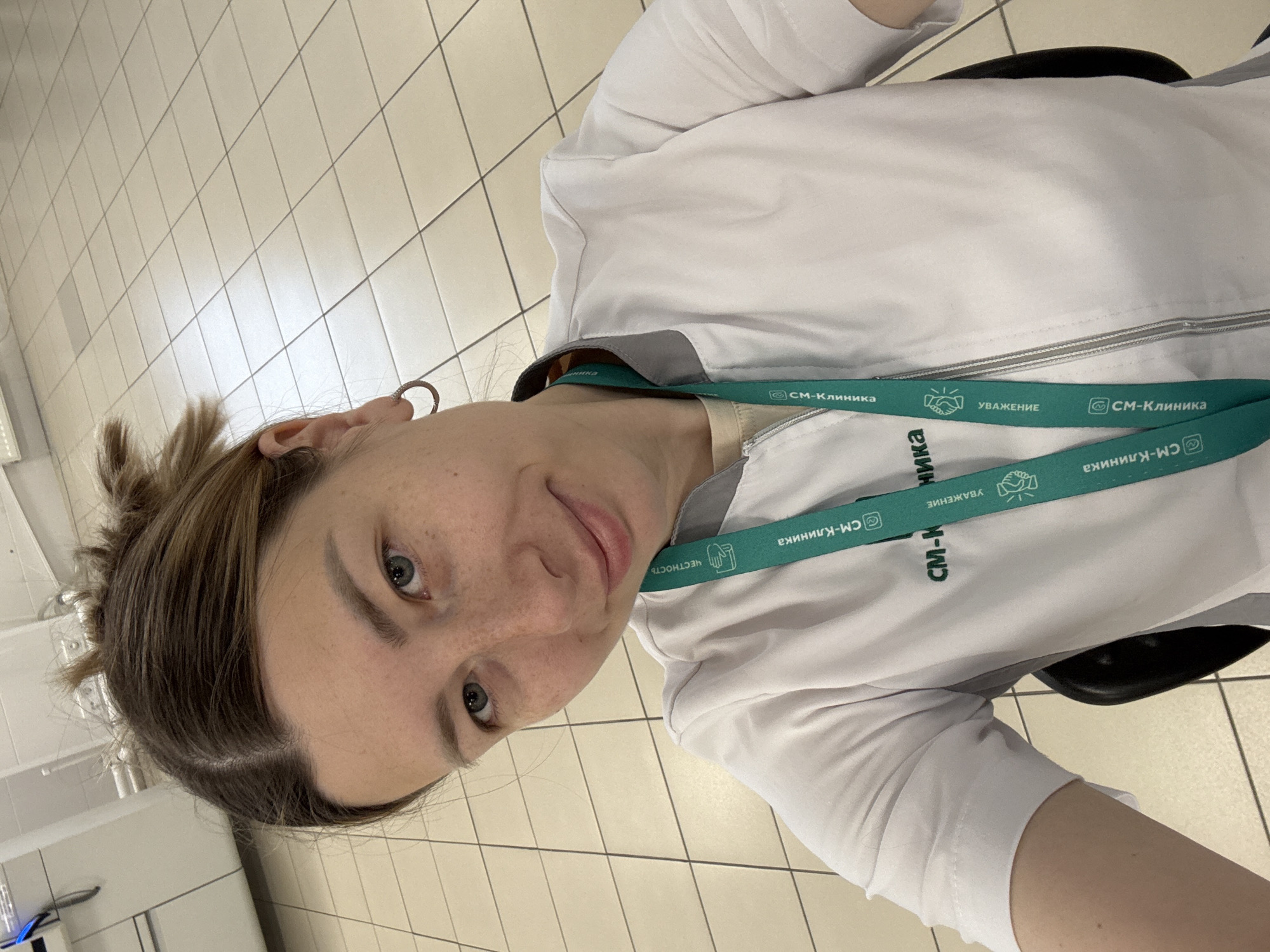 A young woman wearing a white medical coat and a lanyard that identifies her as working at "SM-Klinika" (СМ-Клиника) is in what appears to be a clinic. The lanyard also contains text in Russian, which translates to “Honesty” and “Respect.” The location is likely in Moscow, Russia, since SM-Klinika is a medical network with locations in the city. She has light hair and is wearing a hoop earring. The background shows light-colored square floor tiles. She appears to be taking a selfie.
