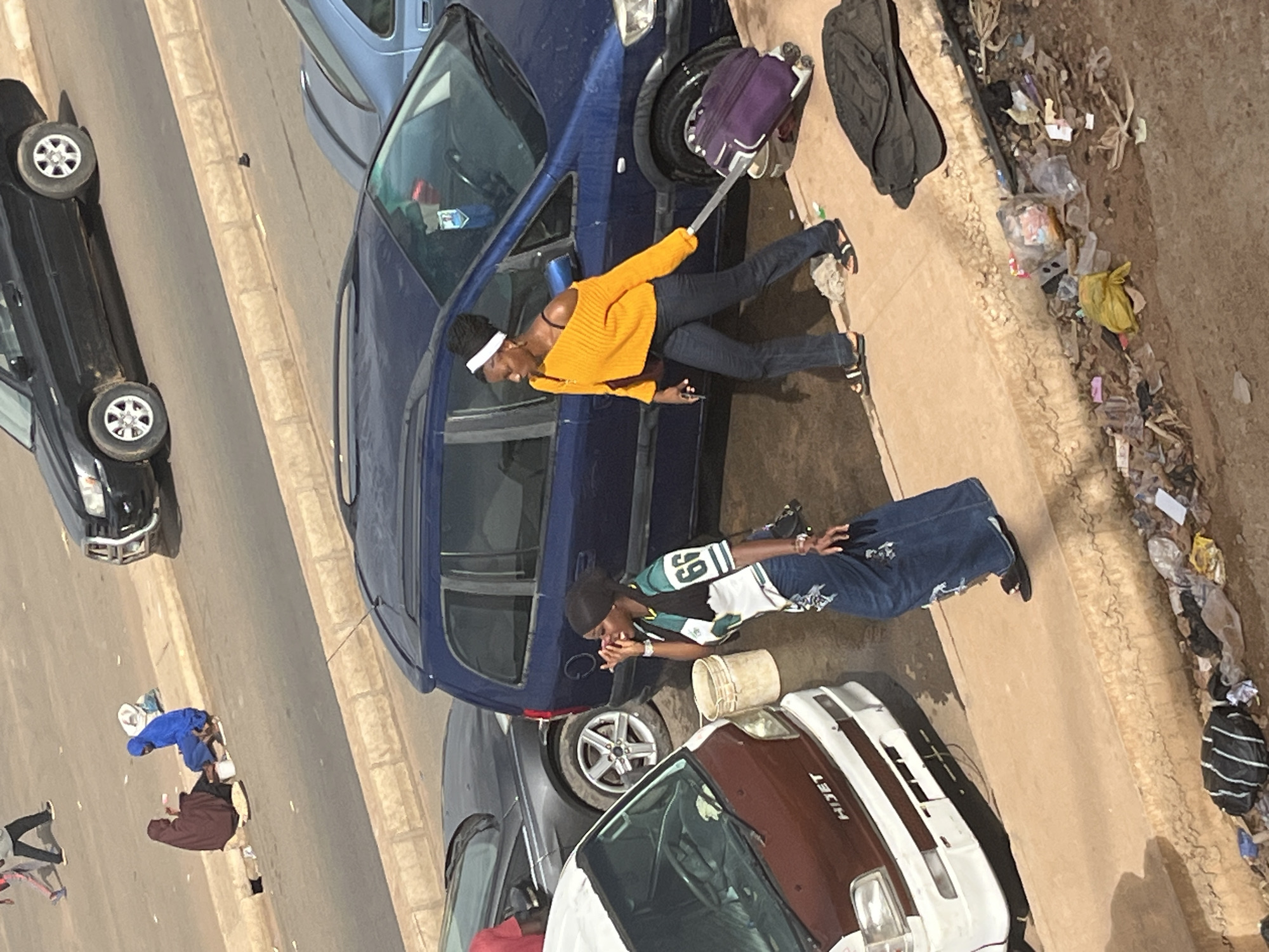 This image captures a scene on a dusty street in Jos, Nigeria. Two individuals are the primary subjects. One, a person wearing an orange sweater and dark pants, is standing near a blue car, holding a selfie stick with a purple bag attached. The other person, dressed in a green and white shirt and dark jeans, is seated next to a white bucket, seemingly engaged in a conversation or resting.
The setting appears to be an outdoor environment, possibly a street or roadside, with several cars present. A black car is parked on the left side of the image, and a blue vehicle is prominently featured. Another vehicle, a white and maroon van with "HIJET" visible on the side, is parked on the right, facing away from the viewer. The ground is a mix of dirt and paved surfaces, with scattered debris, including plastic bags and other trash, along the right edge of the frame, suggesting an urban or semi-urban environment.
The lighting suggests it is daytime, with the sun casting shadows, indicating it could be mid-morning or late afternoon. The overall atmosphere appears casual, with the individuals appearing relaxed. There are no overt signs of distress or urgency. The scene depicts a mundane moment of everyday life, possibly involving travel or waiting, with the selfie stick hinting at the potential for social media activity. The presence of trash indicates potential sanitation challenges in the area.