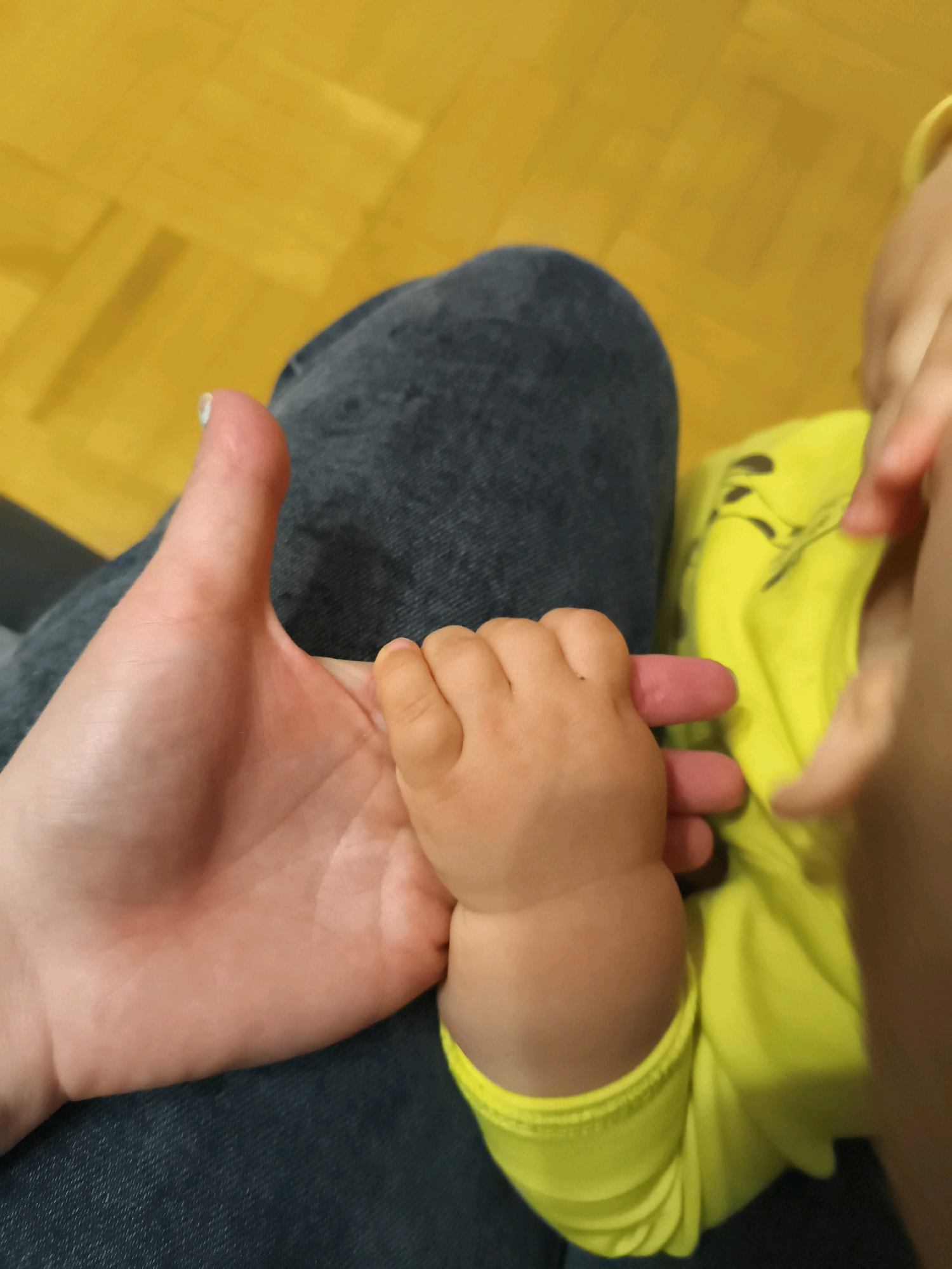 In an overhead shot, an adult's hand and a baby's hand are clasped together. The adult's hand is on the left, with fingers spread slightly. The baby's hand is smaller and appears to be holding one of the adult's fingers. Both hands are positioned over a person's lap, clad in dark denim jeans. To the right of the hands, a bright yellow garment, possibly a shirt, is visible. The background is a yellow floor, possibly linoleum or wood, with a visible seam pattern. The scene is brightly lit, likely by an overhead light source.