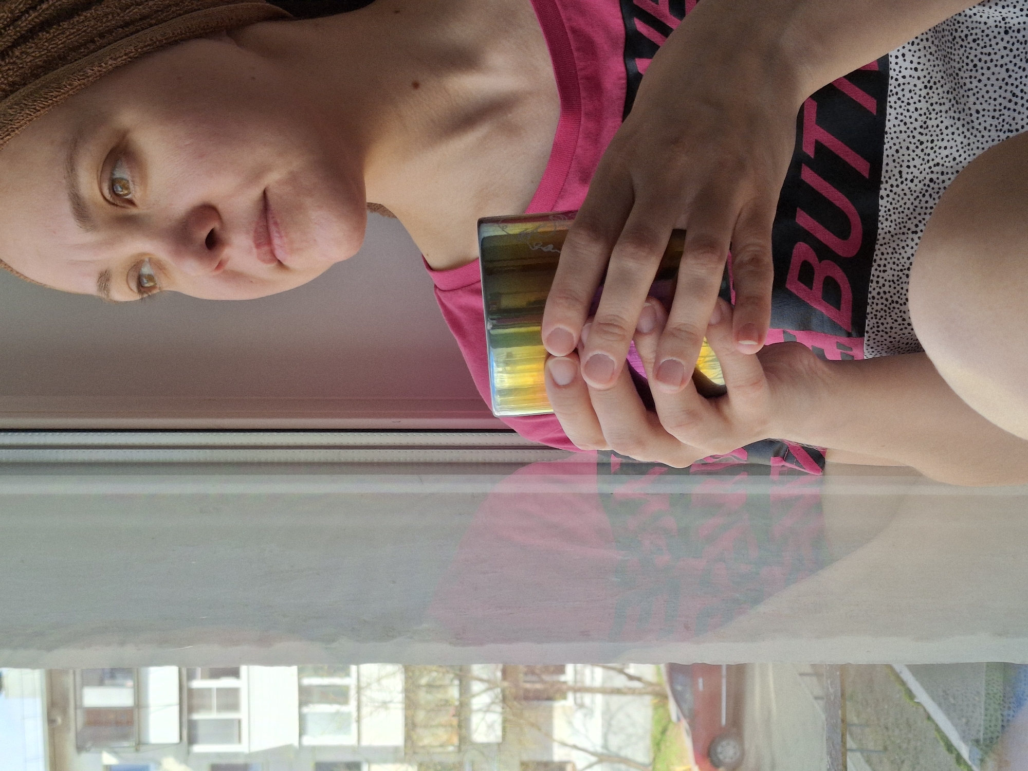 An eye-level, rotated shot captures a light-skinned person, appearing female, leaning forward indoors, looking towards the camera with a neutral to slightly contemplative expression. They have warm brown eyes and natural eyebrows, and their head is covered with a light brown textured wrap or towel. The person is wearing a vibrant pink t-shirt with a black pattern, and a portion of a white fabric with small black polka dots (likely shorts or a skirt) is visible on the right.
The person holds a multi-colored, iridescent glass in both hands, resting it on their lap or a surface. Their fingers are visible around the glass, which reflects various hues.
The scene is set indoors next to a window, through which the outside world is reflected. The bottom portion of the image shows a clear reflection of the person's upper body, including their pink shirt and the patterns on it. Beyond the reflection, through the window, an urban or suburban landscape is visible, featuring multi-story buildings, some bare trees, and a red car parked on the street. The natural lighting suggests it is daytime with clear weather.
On the person's pink t-shirt, a black pattern includes partially visible text; the word "BUT" is clearly legible. The location context for this image is Chaikovsky, Russia.
