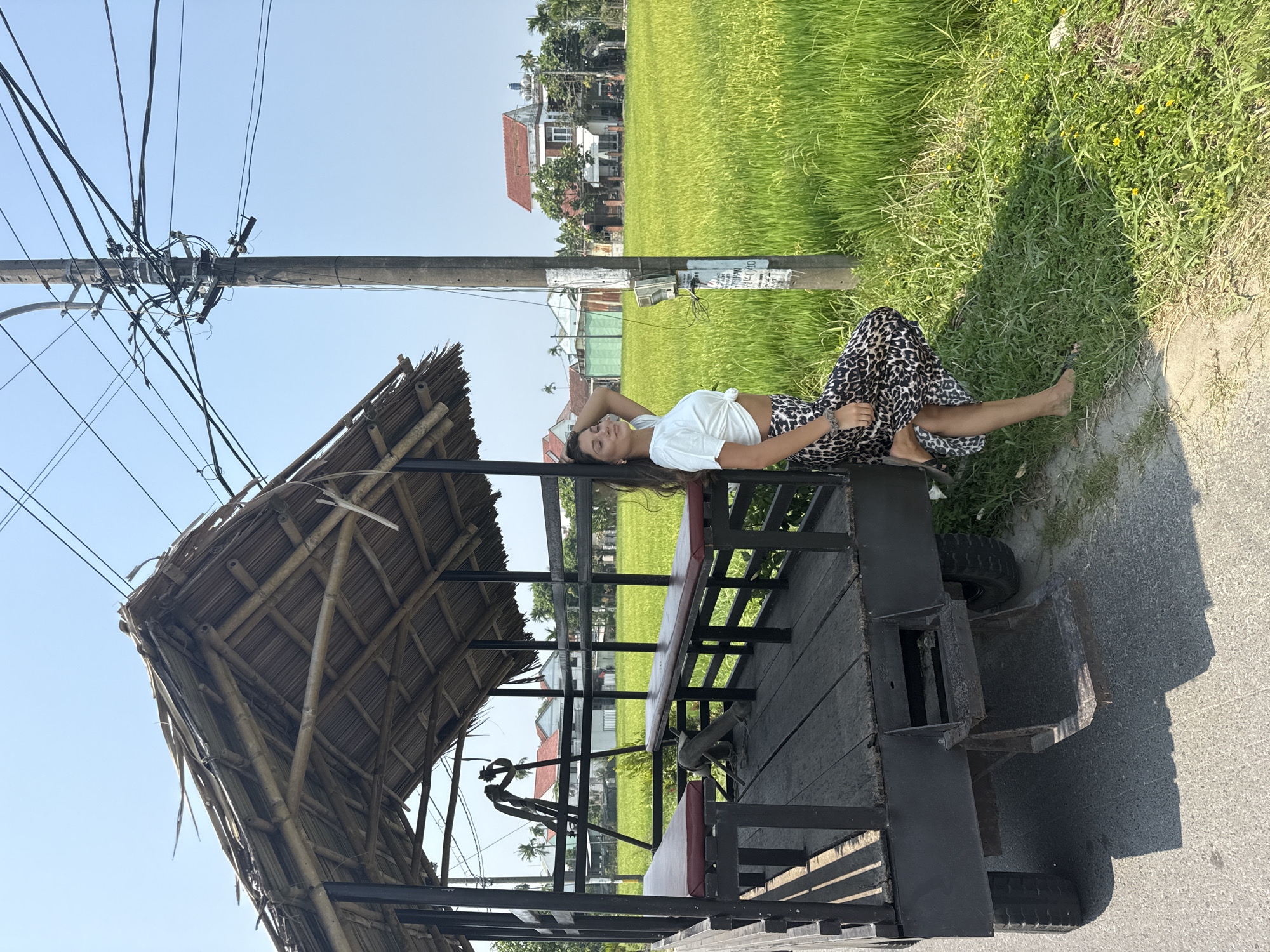 A horizontal medium shot depicts a woman lounging on a vehicle adjacent to a rice paddy in Đà Nẵng, Vietnam. She is fair-skinned, dark-haired, and wears a white t-shirt and animal-print pants, her feet are bare. She lies on her left side with her left arm raised, head resting on her hand. The vehicle appears to be a wooden platform with a bamboo roof and black metal supports, possibly a modified trailer or cart. It sits on a paved road bordering a field of tall, green rice plants. A utility pole with numerous wires extends into the upper left of the frame against a clear blue sky. In the background, there are houses with red-tiled roofs. The overall lighting is bright, suggesting a sunny day. The scene is a mix of rural and urban elements.