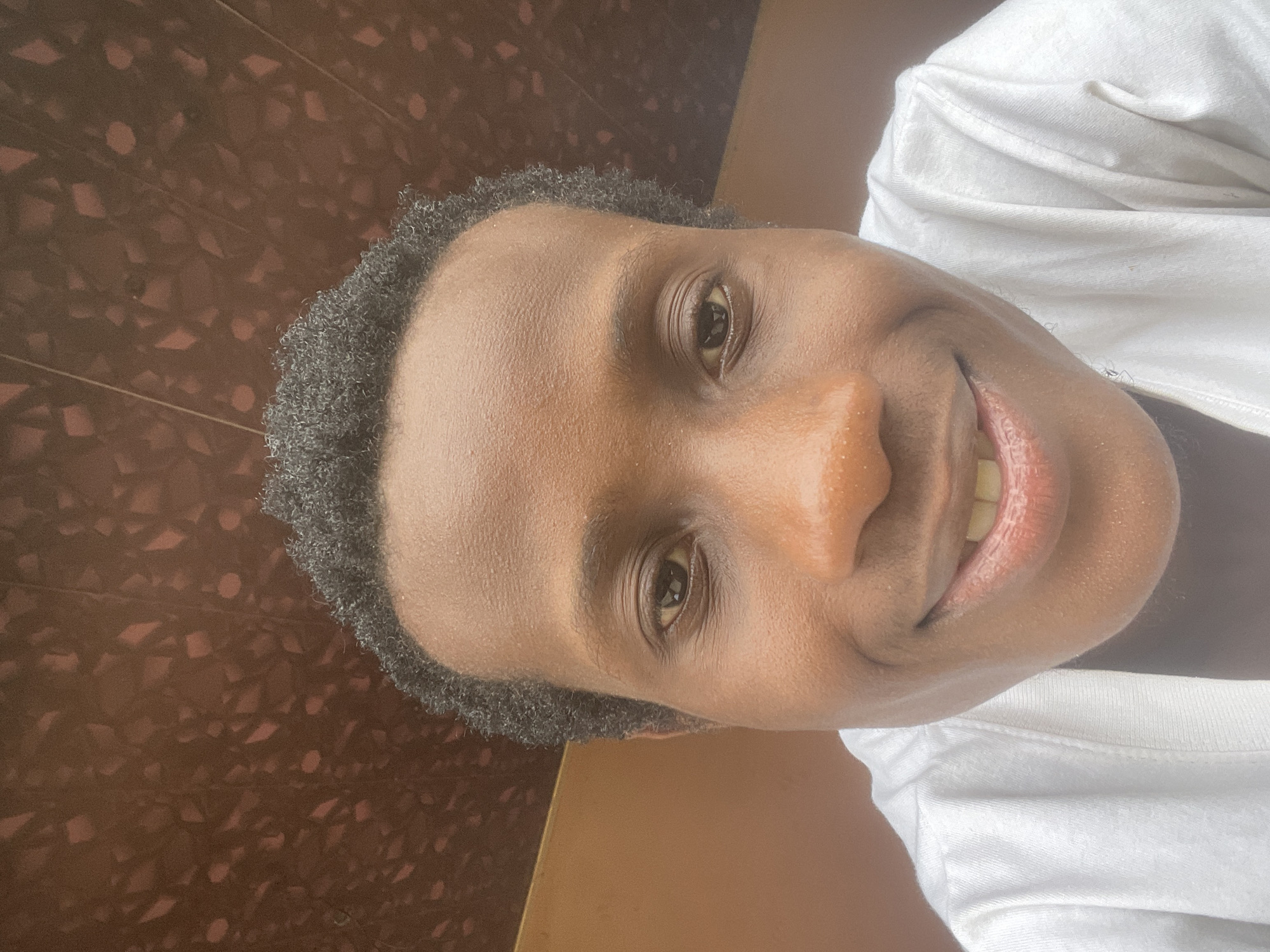 A low-angle, close-up, selfie-style shot shows a young Black person with short, dark, curly hair, dark eyes, and brown skin. The person is wearing a white t-shirt and smiling, revealing their teeth. The background consists of a brown, textured ceiling and a tan wall. This photograph was taken in Ihiagwa, Nigeria.