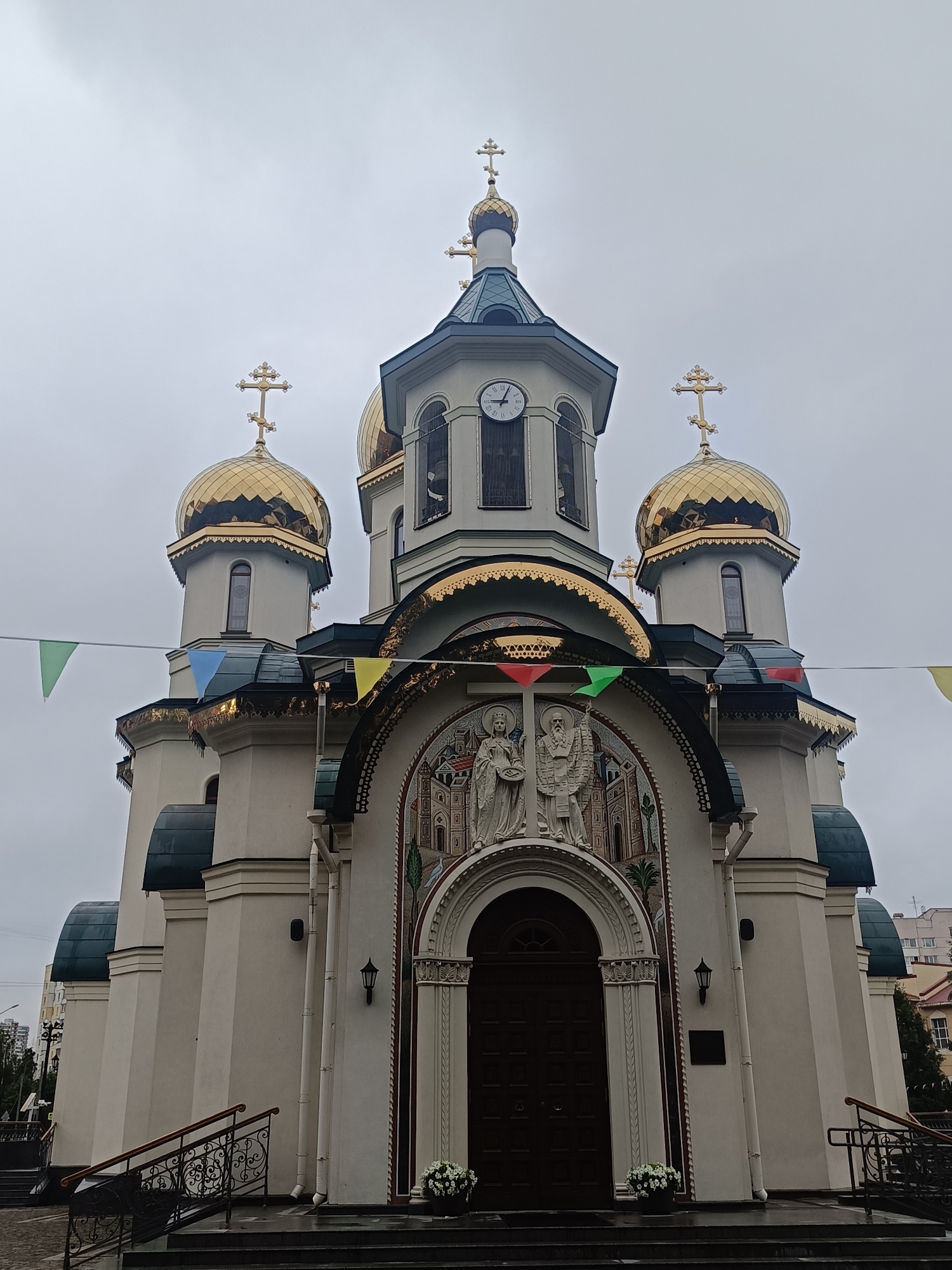 The image shows a low-angle exterior view of the Church of the Resurrection of Christ in St. Petersburg, Russia, under an overcast sky. The church is an Orthodox church with a light-colored facade and multiple gold-domed roofs topped with Orthodox crosses. A large clock face is visible on the central tower. Decorative banners, triangular in shape and in various colors, are strung across the front of the building. Above the main entrance, an arched mosaic depicts religious figures. The entrance itself is a large, dark wooden door. Steps lead up to the entrance, flanked by ornate black metal railings. Planters with white flowers are visible at the base of the entrance. The surrounding environment is only partially visible, with hints of urban buildings in the background.