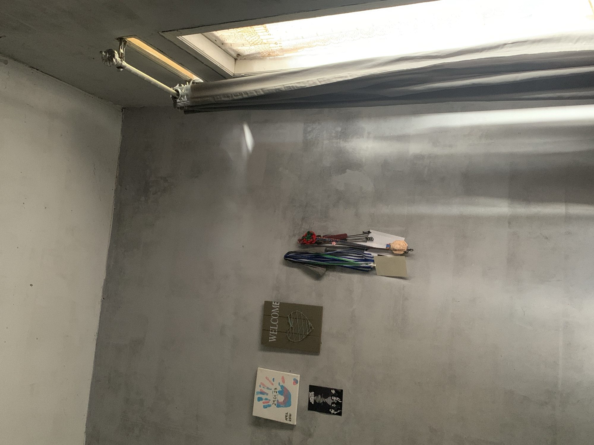 The image depicts an indoor scene, likely a wall in a room, with natural light entering from a window on the right side. The wall appears to be a rough concrete or stucco finish, predominantly gray, with some variations in texture and color.

Decorations are affixed to the wall. On the left, there is a small square framed artwork with colorful handprints and text, including "24.01.23" and "APRIL RIFQI." Below this is a smaller, rectangular black and white image, possibly a band poster, with text that appears to read "ARCTIC MONKEYS." To the right of the handprint artwork is a square plaque with the word "WELCOME" written at the top and a stylized heart made of string in the center. To the right of the "WELCOME" plaque, a collection of ribbons and medals are hanging from the wall, some with bronze-colored discs visible.

A curtain, appearing to be a neutral gray fabric, is drawn partially across the window on the right. The window itself is narrow and appears to have a decorative lace or patterned sheer curtain on the inside. Sunlight streams through the window, casting distinct light patterns and shadows on the wall. The upper part of the image shows the junction of the wall and ceiling, which is also a lighter shade of gray. The window frame is visible, as is a curtain rod and mounting bracket.

There are no people or active interactions visible in the image. The scene is static and appears to be a personal space decorated with mementos. The overall atmosphere is somewhat dim, with the strong contrast between the lit window area and the shadowed wall. The location in the city of Utama, Indonesia, cannot be determined from the visual information provided.