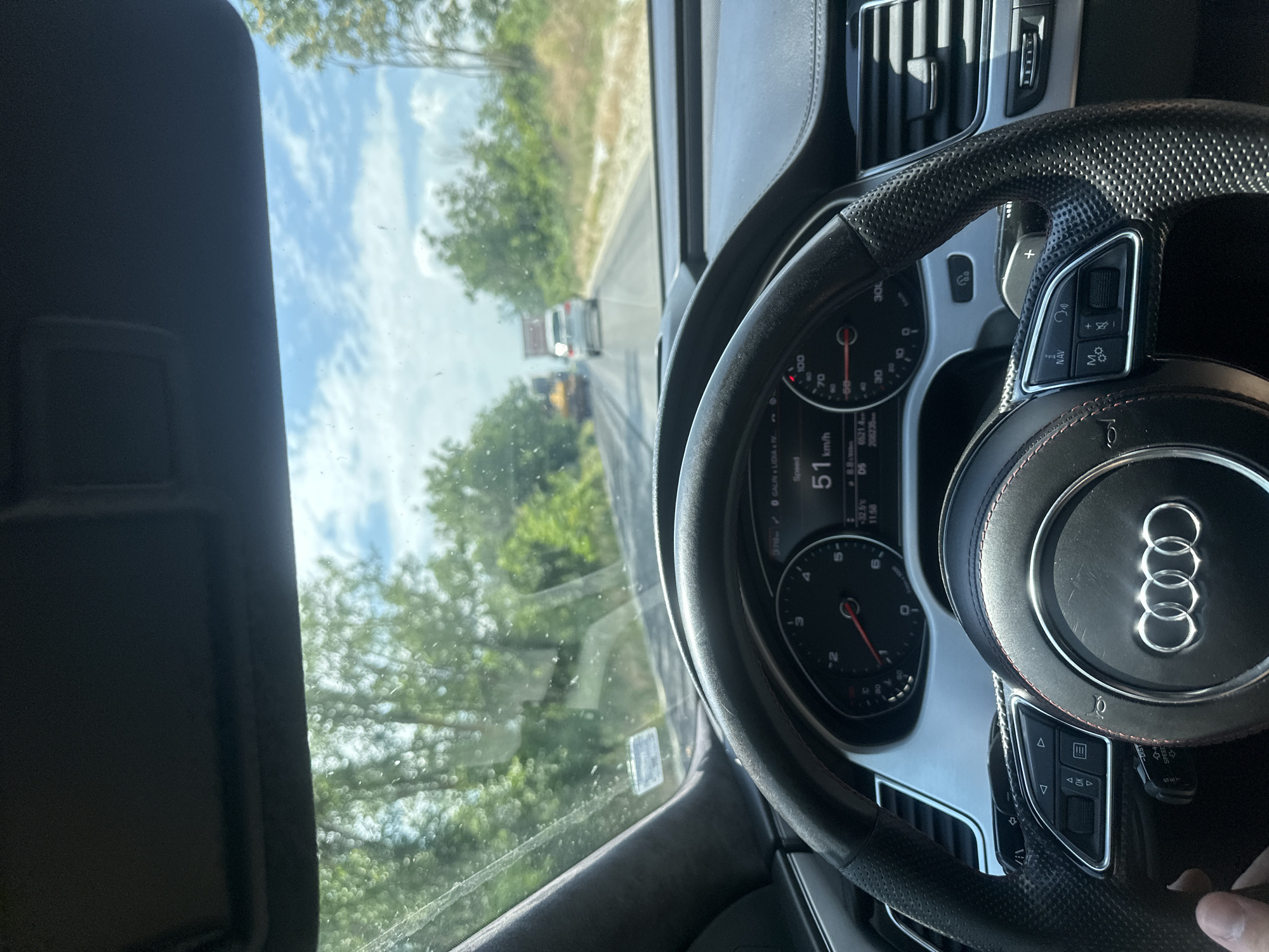 The image is a first-person view from the driver's seat of an Audi car, showing the dashboard and the road ahead. The car is traveling on a two-lane road, with trees and vegetation on either side. The sky is partly cloudy. In the distance, there are other vehicles on the road. The digital display on the dashboard shows the speed as 51 km/h. The scene is consistent with a rural or suburban environment, and the specific mention of Polikraishte, Bulgaria, suggests this location. There are no people clearly visible in the image other than the driver's hand on the steering wheel.