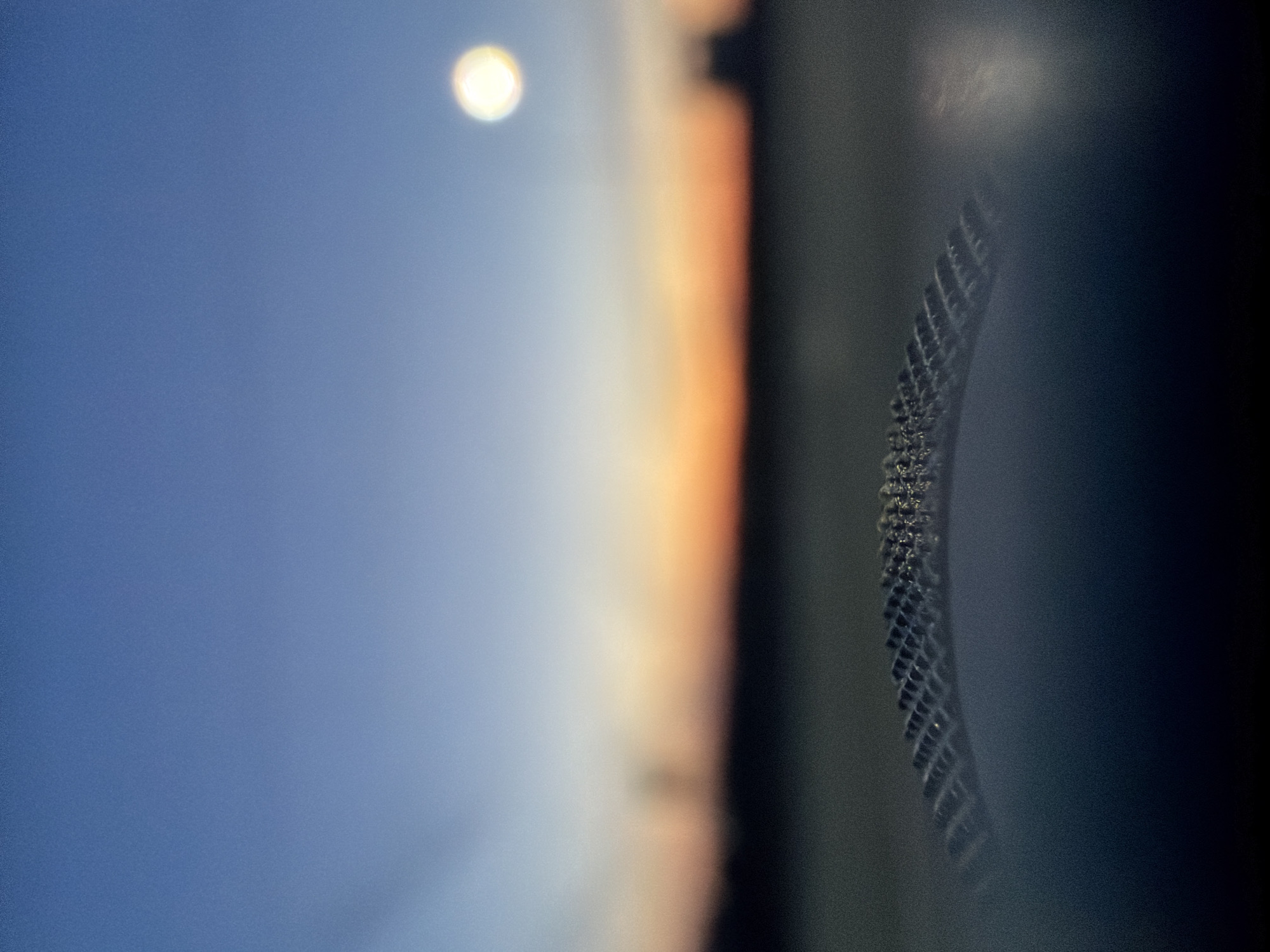 The image appears to be a close-up, possibly a macro shot, focusing on a portion of a microchip or similar electronic component. The component has a dark, possibly black, surface with a grid-like pattern of metallic or reflective squares. These squares seem to be arranged along a curved edge. The background is out of focus, showing a gradient of blue and orange, suggesting a sky with a light source (possibly the sun or moon) blurred in the upper-left corner. No people, specific actions, or interactions are directly visible in the frame. The image does not contain explicit information or references that would tie it to the city of Kuregovo, Russia. The environment is defined by the component's surface and the blurred background light.