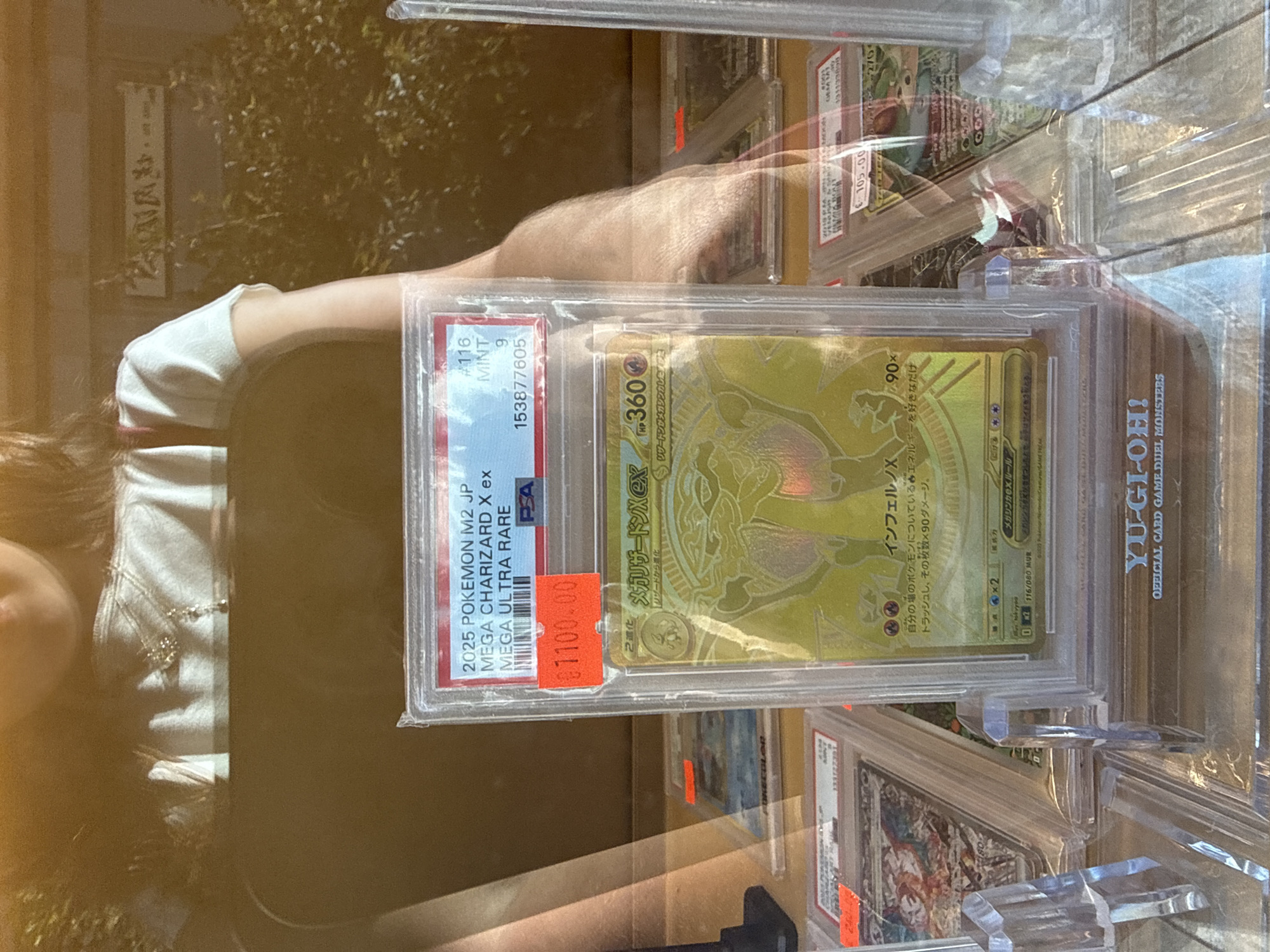 A person, partially visible from the chest up, wearing a white t-shirt and a necklace, is standing behind a display of trading cards. The foreground shows several graded trading cards in protective cases. The most prominent card is a gold-colored Pokemon card, identified as "2025 POKEMON M2 JP MEGA CHARIZARD X ex MEGA ULTRA RARE" with a PSA grade of 9 and a serial number 153877605. It has a red price sticker showing "$1100.00". Other graded cards are visible behind and to the sides, some with price stickers. The display appears to be at an outdoor market or store, with foliage and a blurred background suggesting trees or bushes. The overall lighting suggests it is daytime. The location is specified as Milan, Italy.