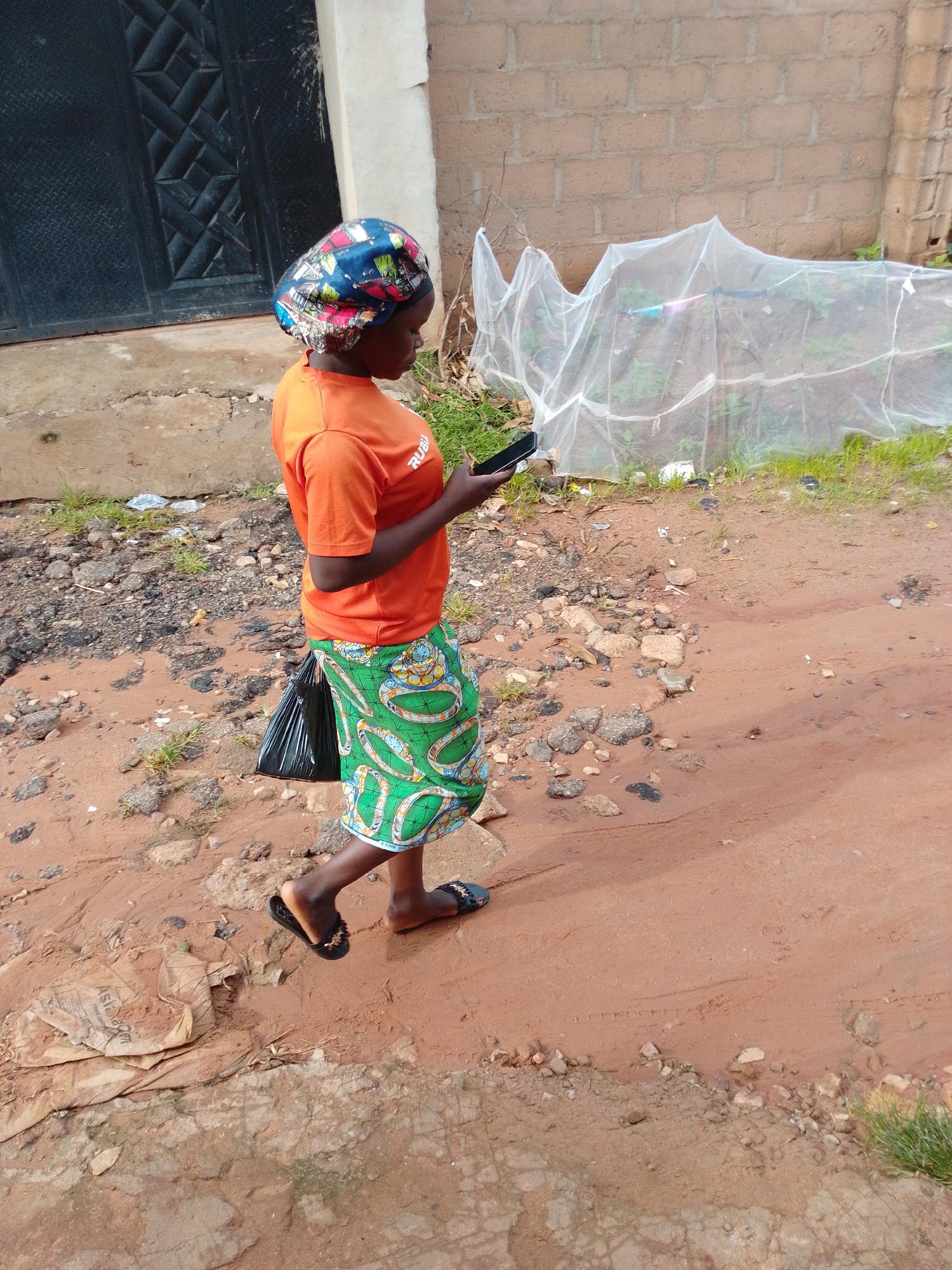 A woman wearing an orange t-shirt and a colorful headscarf walks on a dirt path in Gombe, Nigeria. She is holding a black smartphone in her right hand and appears to be looking at it. A black plastic bag is tucked into the waistband of her green and white patterned skirt. The ground is a mix of red dirt, gravel, and some scattered rocks. In the background, there is a brick wall and a white mesh covering what appears to be a garden bed. A dark metal door is visible to the left of the frame. The overall environment suggests an outdoor, possibly semi-rural or informal urban setting.