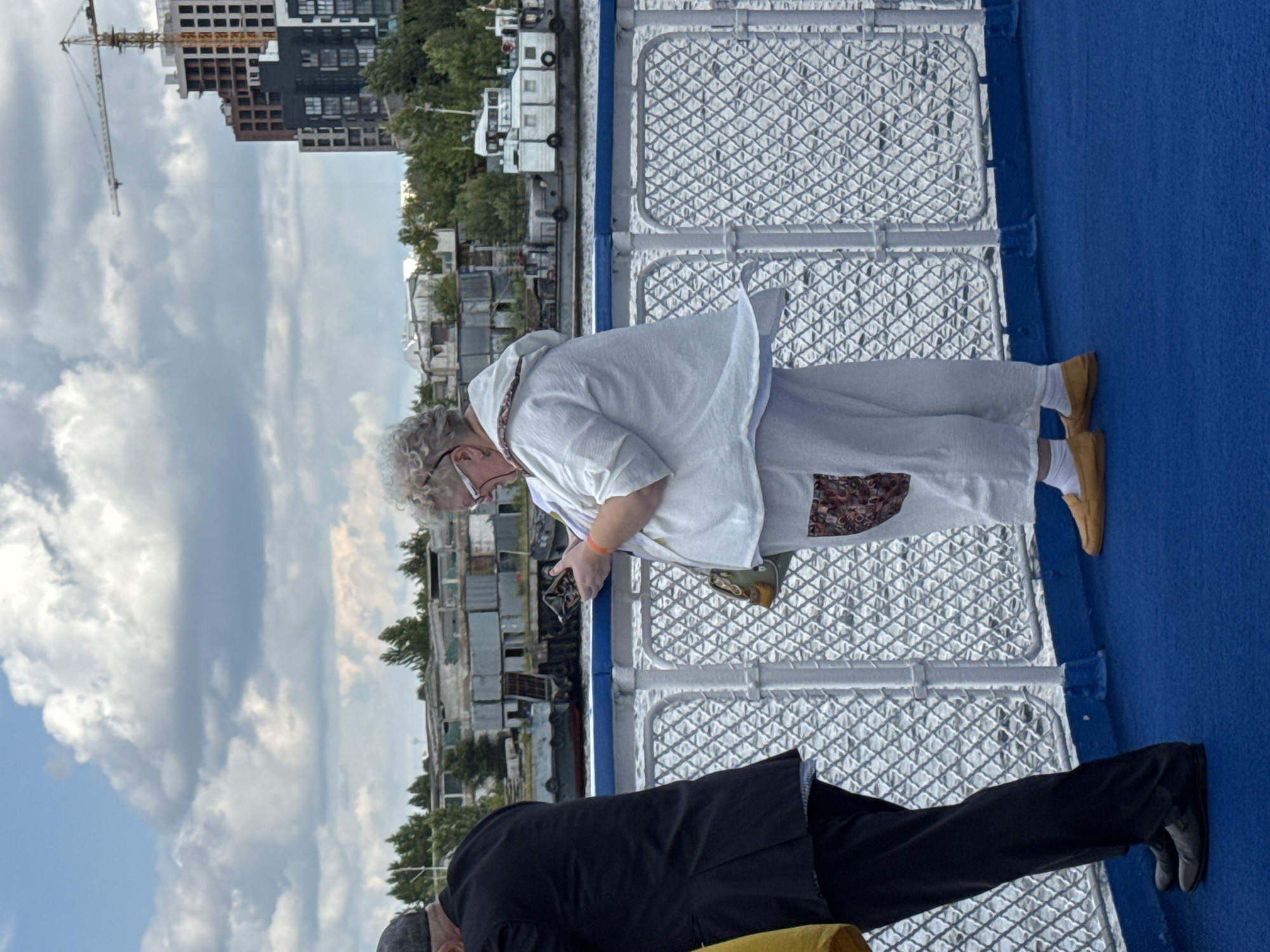 On a bright, cloudy day, two individuals stand on the deck of a vessel, likely a ferry, with a blue deck and a white railing. In the foreground, a woman with curly gray hair and glasses is looking at her phone. She is wearing a white hooded jacket and white pants, with a patterned detail on her right leg. She has a green and tan purse slung over her shoulder. To her left, a man in a black suit and dark cap is partially visible, also looking down. In the background, across a body of water, a cityscape is visible. This includes a crane, partially constructed buildings, and other industrial structures, suggesting a developing urban area. The presence of buildings under construction and a crane is indicative of the city of Kazan, Russia. The water appears choppy, reflecting the sky. The railing has a diamond-shaped mesh pattern.