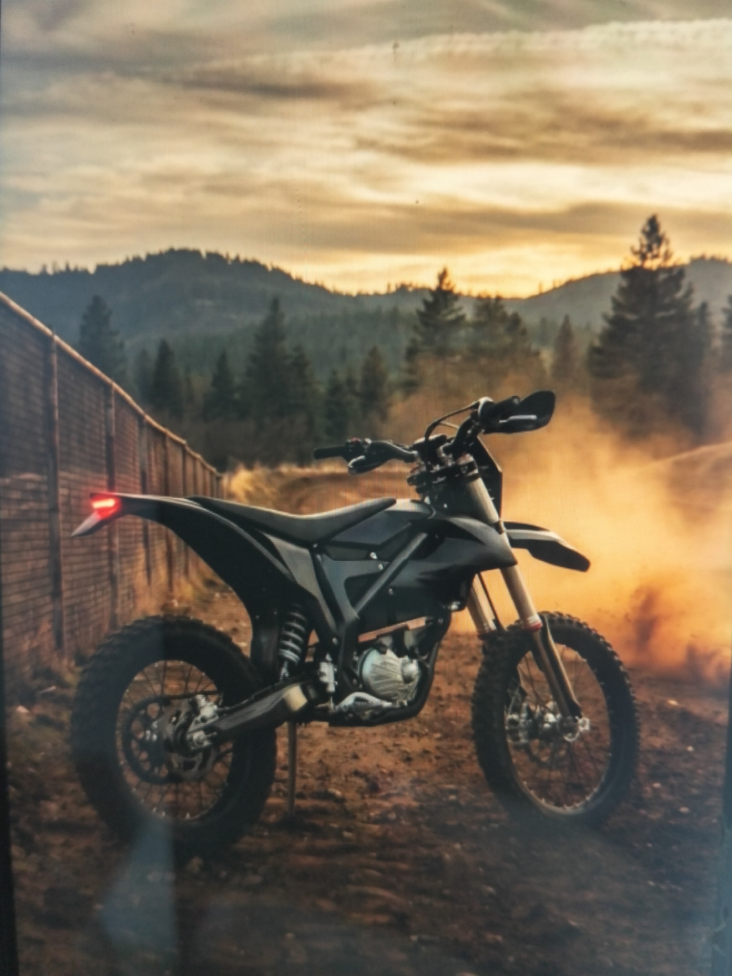 The image shows a black electric dirt bike parked on a dirt road next to a wooden fence. The bike is the primary subject, positioned in the center of the frame. It features knobby tires, a suspension system, and a visible motor component. The background consists of a forested hillside under a cloudy, sunset sky. Dust is kicked up in the air behind the bike, suggesting recent movement. The overall lighting is warm and atmospheric, indicative of late afternoon or early evening. There are no people present in the image. The location is not specified as being in Payagaboh, Indonesia.
