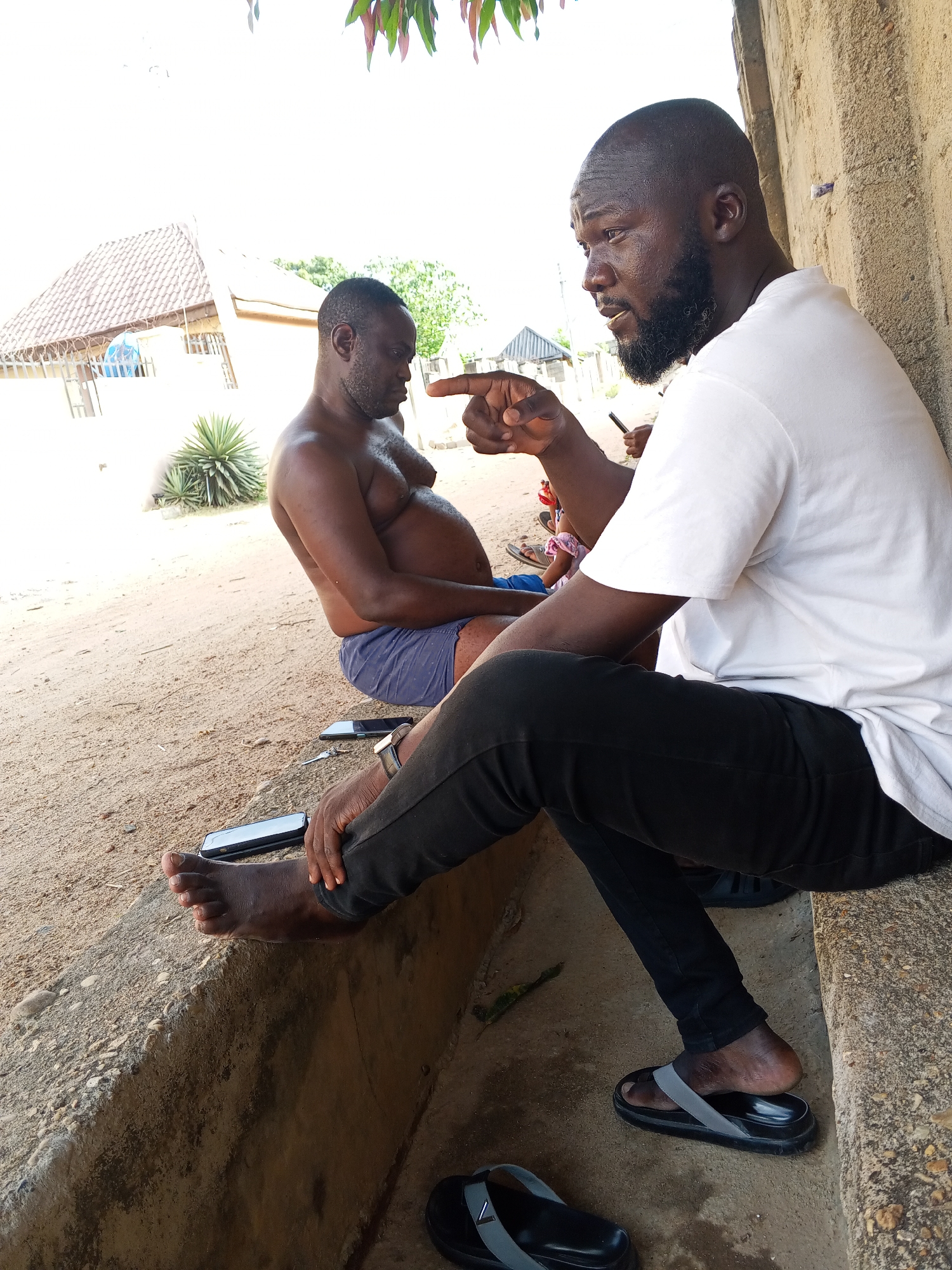 The image captures two men in an outdoor, sunlit setting in what appears to be Adonkolo, Nigeria.
In the foreground, to the right, a dark-skinned man with a bald head and a short beard is seated on a low, weathered concrete ledge. He wears a white t-shirt and black trousers. His right arm is raised, and he is pointing his index finger forward and slightly to the left, with a focused, serious expression, suggesting he is explaining or emphasizing a point. His left foot, bare, is resting on his right leg, while a black sandal with a grey strap is on his left foot. Another matching sandal lies on the ground beneath him.
To the left and slightly behind him, another dark-skinned man is also seated on the same concrete structure. He is shirtless, revealing a prominent belly, and wears blue shorts. He is oriented slightly away from the camera, looking down or to his left, appearing to be listening or in contemplation.
Several personal items are scattered on the sandy, light brown ground between the men. At least three smartphones are visible, two lying flat on the ground and one closer to the shirtless man. A set of keys is also visible on the ground.
The background shows a simple, dry outdoor environment. To the left, part of a building with a reddish-brown tiled roof can be seen under a bright, clear sky. A spiky, agave-like plant grows in the middle ground. The overall lighting suggests it is midday or early afternoon, with bright, direct sunlight indicating warm weather. There is no visible text in the image.