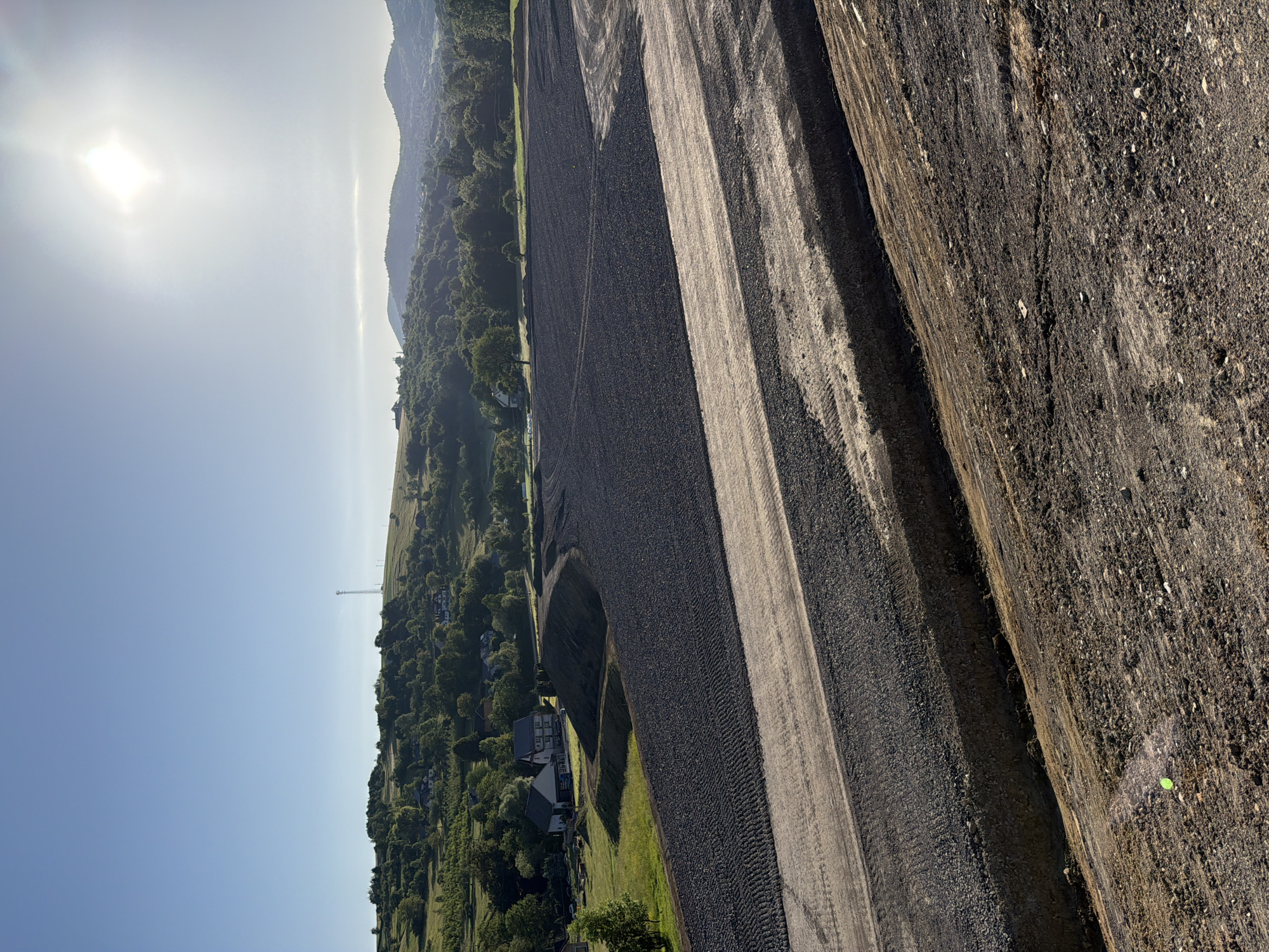 The image depicts an expansive construction site under a bright, hazy sun, suggesting an early morning or late afternoon. The foreground is dominated by raw earth and gravel, with visible tire tracks indicating recent activity by heavy machinery. A series of layered, compacted gravel paths run diagonally across the frame, creating distinct terraces. In the mid-ground, a large, dark, uneven surface, possibly compacted soil or asphalt, stretches out, bordered by more gravel layers. To the left, a small cluster of houses with pitched roofs sits on a green, rolling hillside dotted with trees. In the distance, a telecommunications tower is visible on the crest of a hill, and beyond that, a range of tree-covered mountains under a pale sky. The overall impression is one of a large-scale earthmoving project in a rural or semi-rural setting, specifically identified as being in the vicinity of Stróża, Poland.