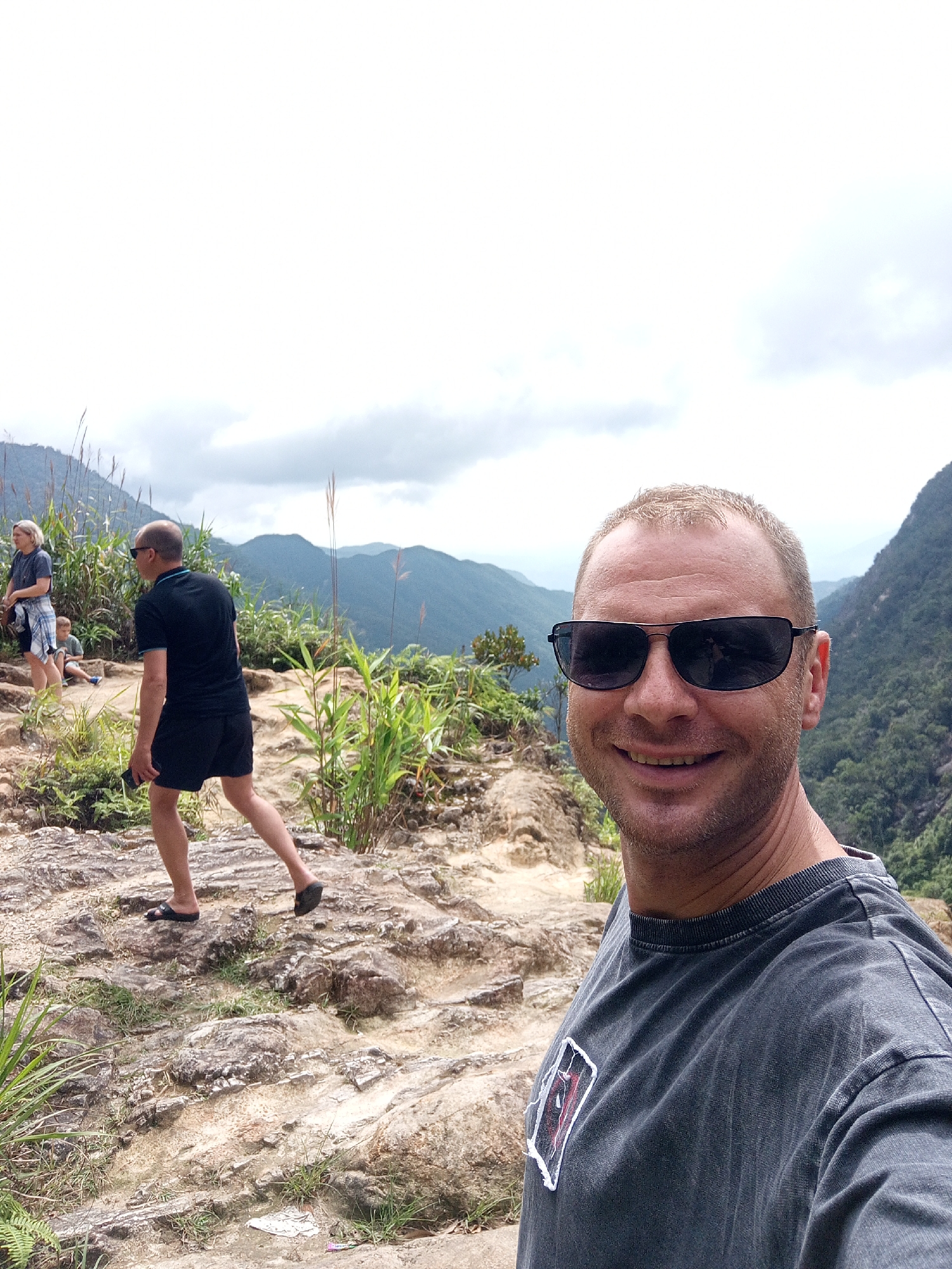 The image shows a group of people on a rocky, mountainous trail. In the foreground, a man with short blonde hair and sunglasses smiles at the camera, wearing a dark gray t-shirt. Behind him, to the left, a man in a black t-shirt and black shorts walks away from the camera. Further back, a woman with blonde hair and a blue and white plaid shirt is visible, along with a child sitting on the ground. The background features lush green mountains and a cloudy sky. The terrain appears to be a hiking trail, likely in a natural, outdoor setting with significant elevation changes. While the image captures a scenic mountainous landscape, there is no specific information or visual cue within the image to confirm its location in Nam Khanh Vinh, Vietnam.