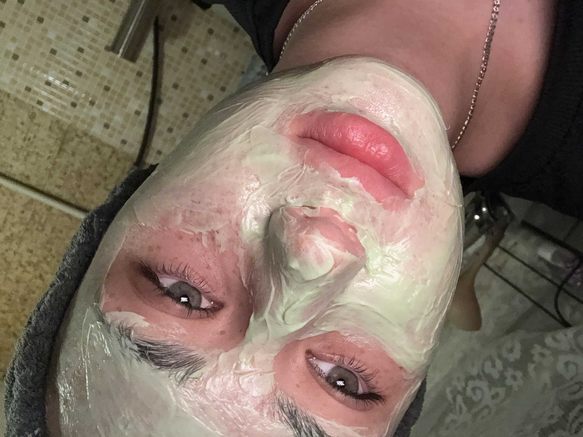 The image is a close-up, low-angle shot of a person's face covered in a pale green face mask, with a dark gray towel wrapped around their head. The person's eyes are open and looking towards the camera. The skin around the eyes and cheeks appears red. The lips are a bright pink color and are slightly parted. A thin silver chain is visible around the person's neck. The background consists of a tiled wall and a patterned cloth. The image is likely taken indoors. There is no information in the image to suggest the location is in the city of Rezh, Russia.