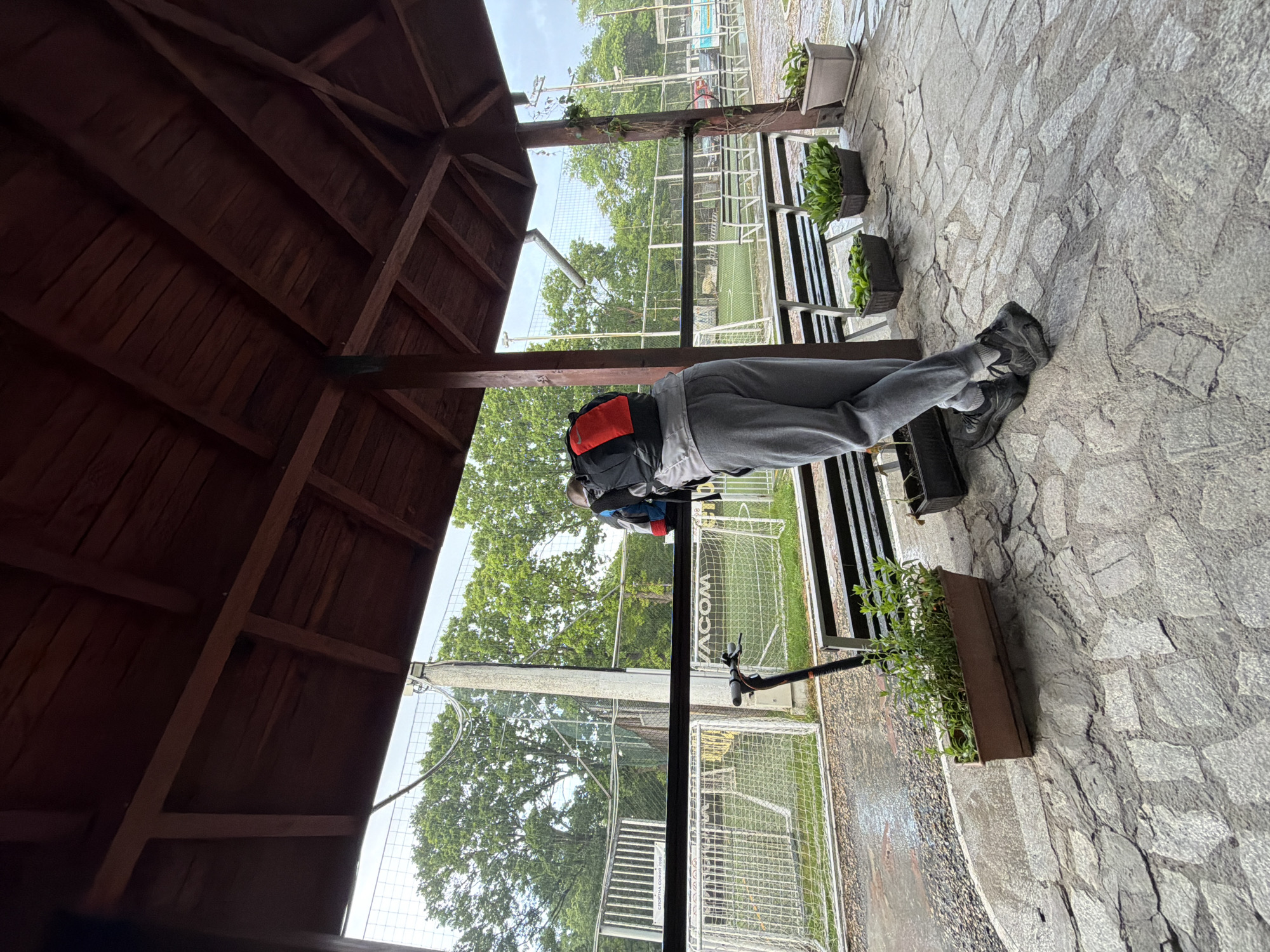 A person wearing gray sweatpants, a gray jacket, and black shoes is sitting on a bench within a wooden structure with a brown roof. The person also has a backpack. Outside the structure is a fenced-in area that looks like a sports field, possibly in Sofia, Bulgaria. A black scooter is adjacent to the bench. Potted plants are nearby on a gray stone patio. The sky is overcast.