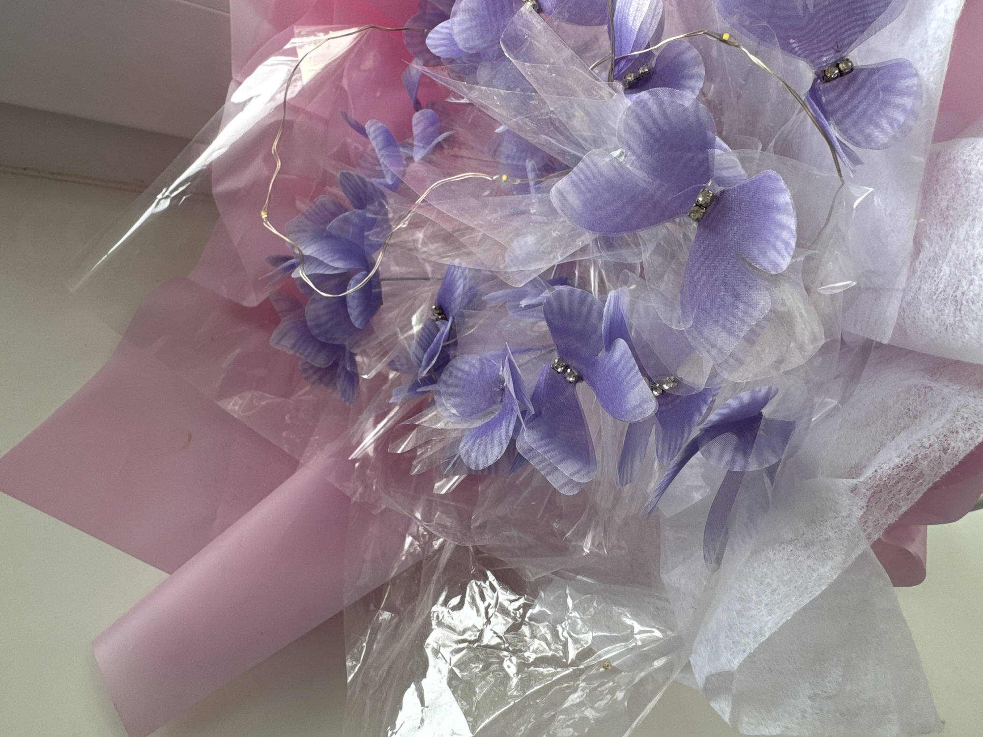 The image shows a close-up, overhead view of a bouquet of artificial lavender-colored butterfly decorations. The butterflies are made of sheer fabric and have small, clear rhinestones at their centers. They are arranged around a bouquet of purple flowers, some of which are also butterfly-shaped. The bouquet is wrapped in layers of pink and clear cellophane, with a layer of white tissue paper at the bottom. Tiny LED fairy lights are woven throughout the bouquet. The background is a plain, light-colored wall. There are no people or animals in the image, and no identifiable landmarks or context that would place this scene in Asha, Russia, or any specific city. The lighting is soft and diffused, suggesting it may be indoors.