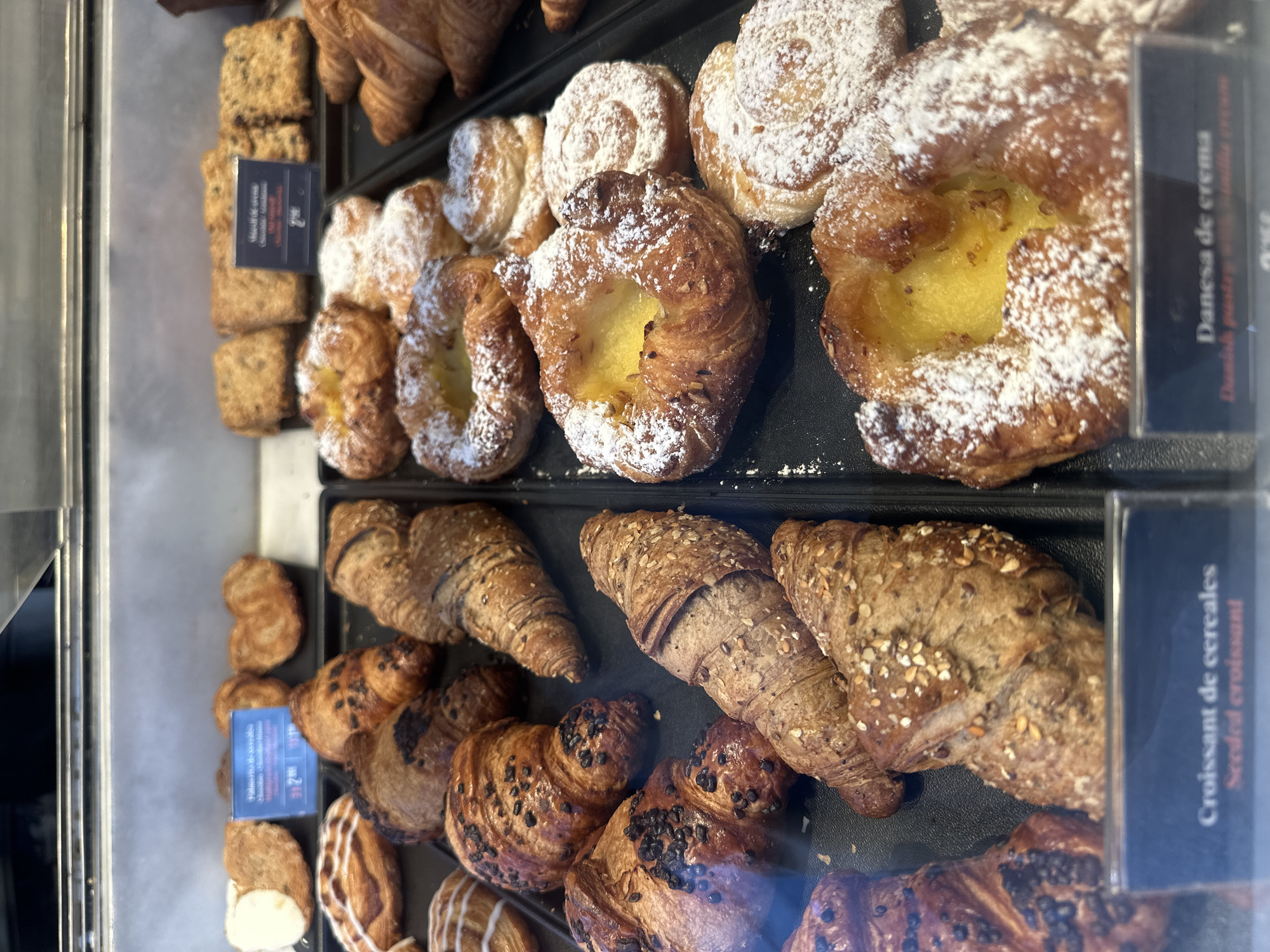 The image displays a vibrant assortment of baked goods meticulously arranged within a glass display case, likely found in a bakery or cafe. The perspective is slightly angled from above, looking down into the brightly lit interior of the case.
In the upper half of the display, several danishes are prominent, characterized by their golden-brown, flaky pastry and a generous dusting of powdered sugar. Many feature a central, bright yellow custard or cream filling. Adjacent to these are rectangular, light brown baked squares, possibly a type of cake or bar, with a coarser texture. A blue label partially visible in this area reads "Danesa de crema" (Spanish for "Cream Danish") and "Danish pastry with vanilla cream," along with a price of "2.90." Another label near the square items has "Maison de..." and "2.90."
The lower half of the display is filled with a variety of croissants and similar pastries. There are standard golden-brown croissants, as well as darker versions seemingly speckled with chocolate pieces. A notable selection includes large croissants covered with a mix of seeds and grains, presenting a rustic, wholesome appearance. A blue label clearly identifies these as "Croissant de cereales" (French for "Cereal Croissant") and "Seeded croissant," also priced at "2.90." Further down, other pastries, some with a white icing drizzle, are visible.
The overall scene suggests an inviting commercial setting, designed to present fresh pastries appealingly to customers. The items are neatly organized on black trays, highlighting their textures and colors. The visible text indicates product names and prices in multiple languages, suggesting a diverse clientele or location.