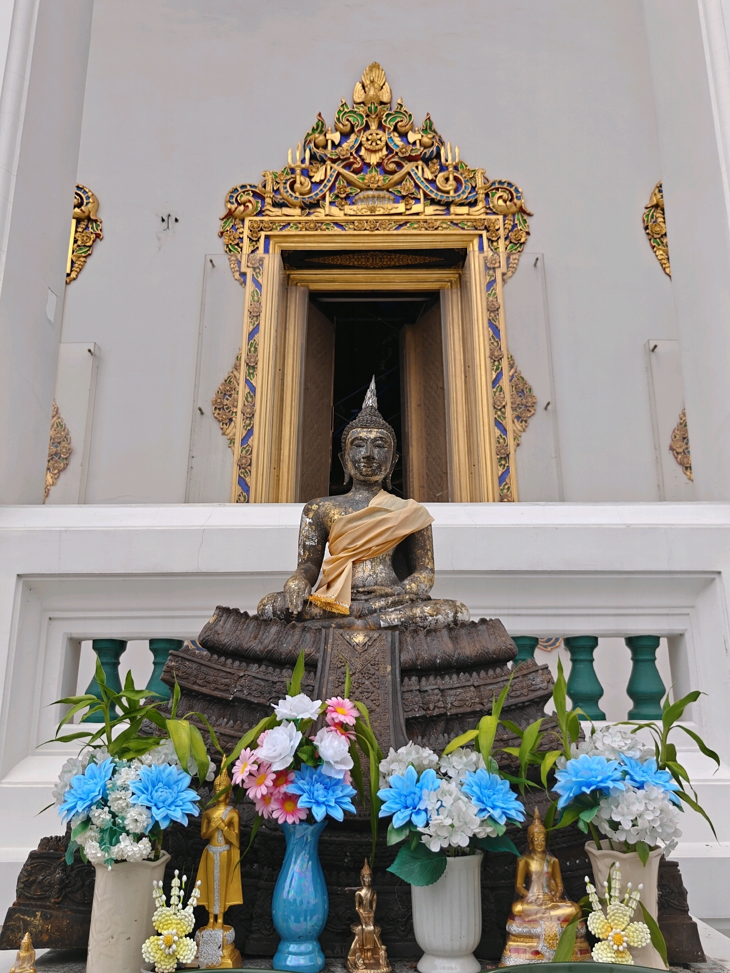 The image shows a weathered, dark-colored Buddha statue seated in front of a white building with ornate golden and blue architectural details above a doorway. The Buddha statue is draped with a gold-colored cloth. In front of the statue, there are several floral arrangements in vases and two smaller golden Buddha figures. The scene appears to be outdoors, possibly in front of a temple or shrine in Bangkok, Thailand.