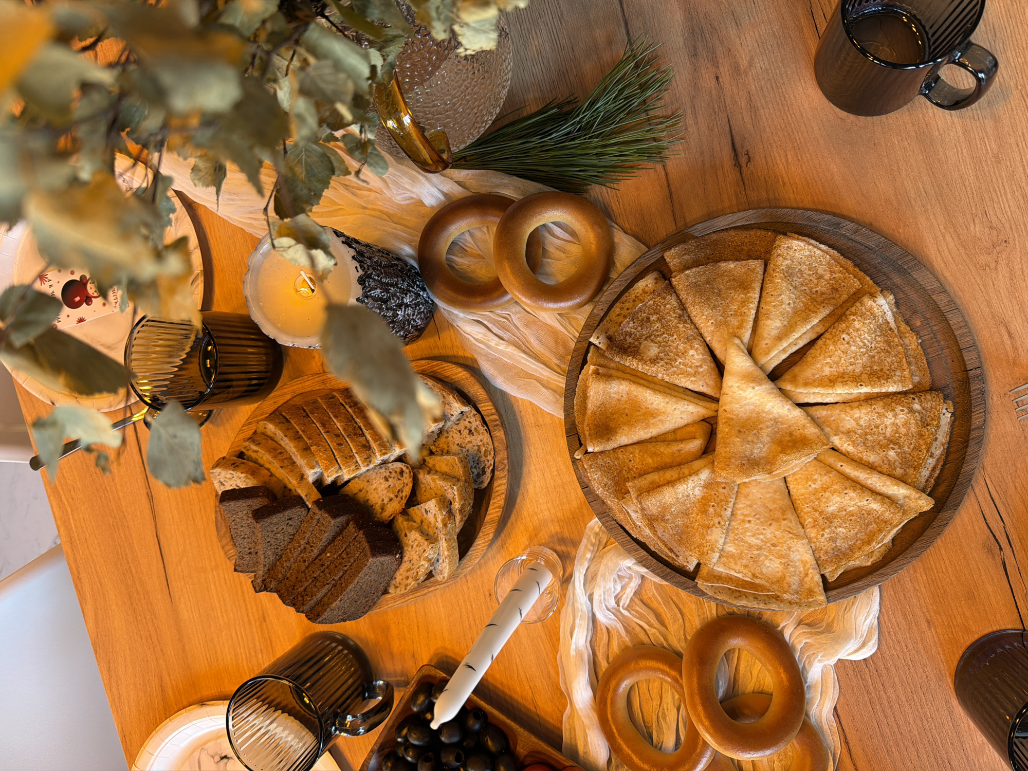 The scene depicts a wooden table laden with various food items. A large round wooden tray holds numerous pancakes, arranged in a circular pattern. Nearby, a smaller wooden tray contains an assortment of sliced bread, including dark and light varieties. Several bagels are positioned near the pancakes and bread. A plate of dark olives is also visible. The table is decorated with a white, textured cloth, adding to the visual appeal. Candles in glass holders and dark-colored glasses are scattered around the table. A vase with greenery is placed on one side, along with a pine sprig. The presence of pancakes, bagels, and olives suggests a possible brunch or celebratory meal. The scene's setting appears to be in Марусино, Russia.