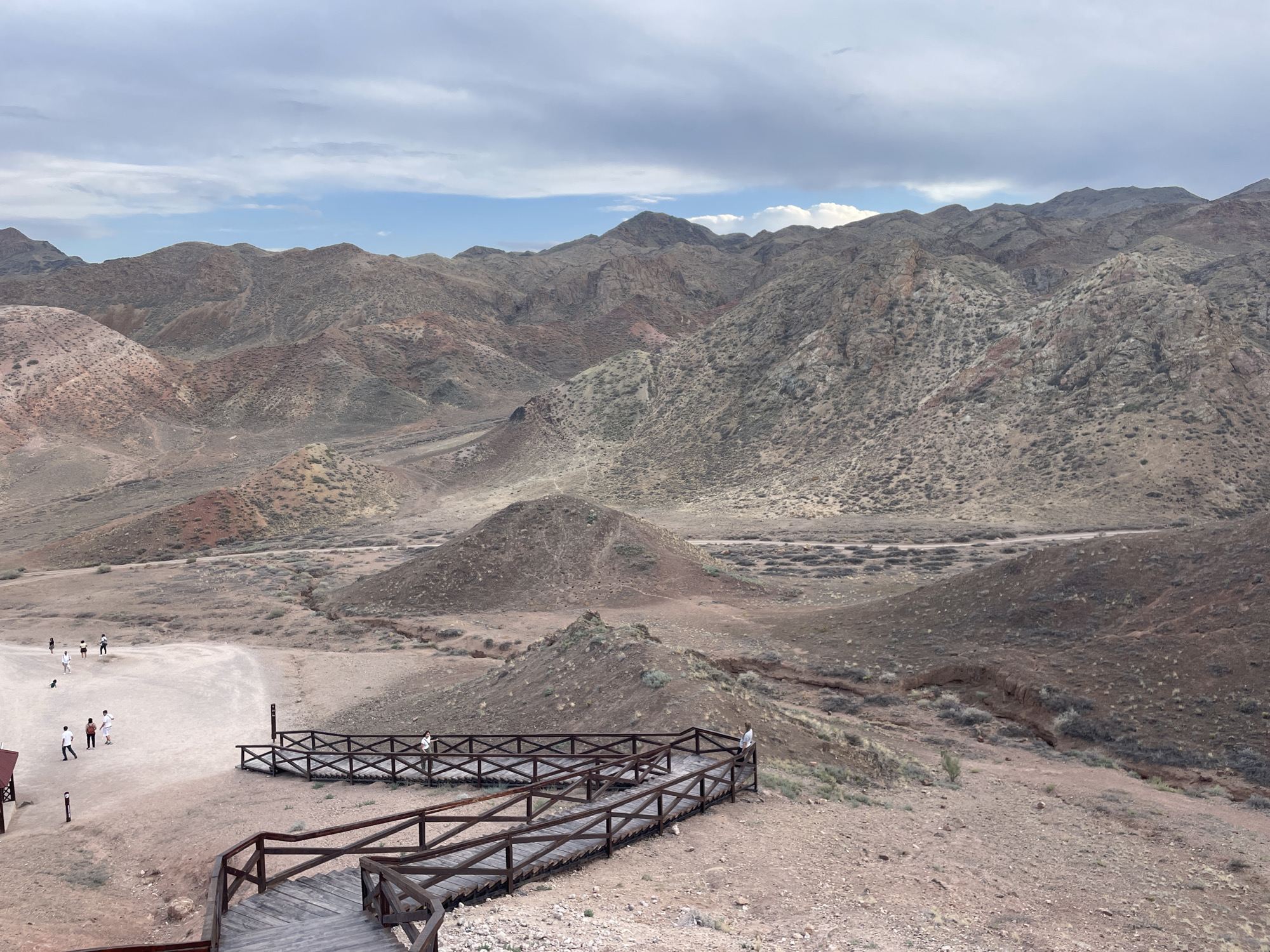 The image depicts a landscape of arid, mountainous terrain with a wooden boardwalk and stairs winding through it. The mountains in the background are characterized by their rocky, eroded surfaces and varied shades of brown, red, and gray. Sparse vegetation, appearing as low-lying bushes and scrub, dots the landscape. In the foreground, a group of people are visible. Several individuals are walking on a dirt path, while others are on the boardwalk. The boardwalk is constructed of dark wood and features railings. The sky is overcast with clouds. The scene is a viewpoint overlooking the mountainous desert landscape, possibly a park or tourist attraction. The location is identified as Tamerlik, Kazakhstan.