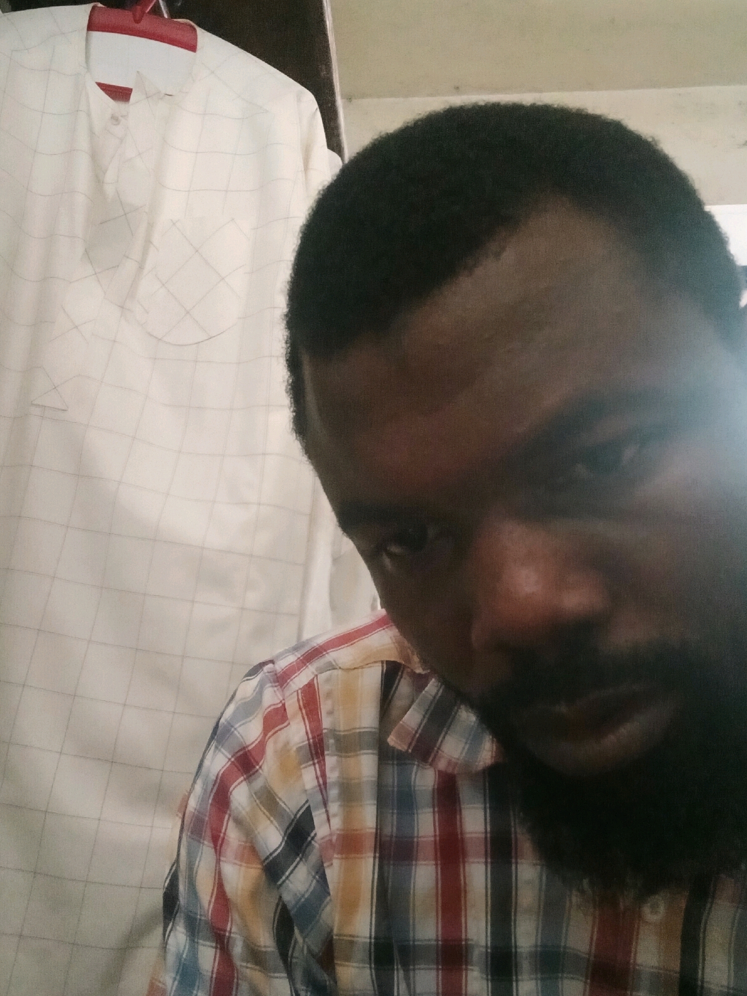 The image shows a close-up, selfie-style shot of a dark-skinned man with a short afro and a beard. He is wearing a red, white, blue, and yellow plaid shirt. In the background, on the left side of the frame, there is a white garment with a subtle grid pattern, possibly a traditional African outfit like an agbada, hanging on a red hanger. The overall lighting suggests an indoor setting. There is no visible indication of the location being in Eko Atlantic, Nigeria.