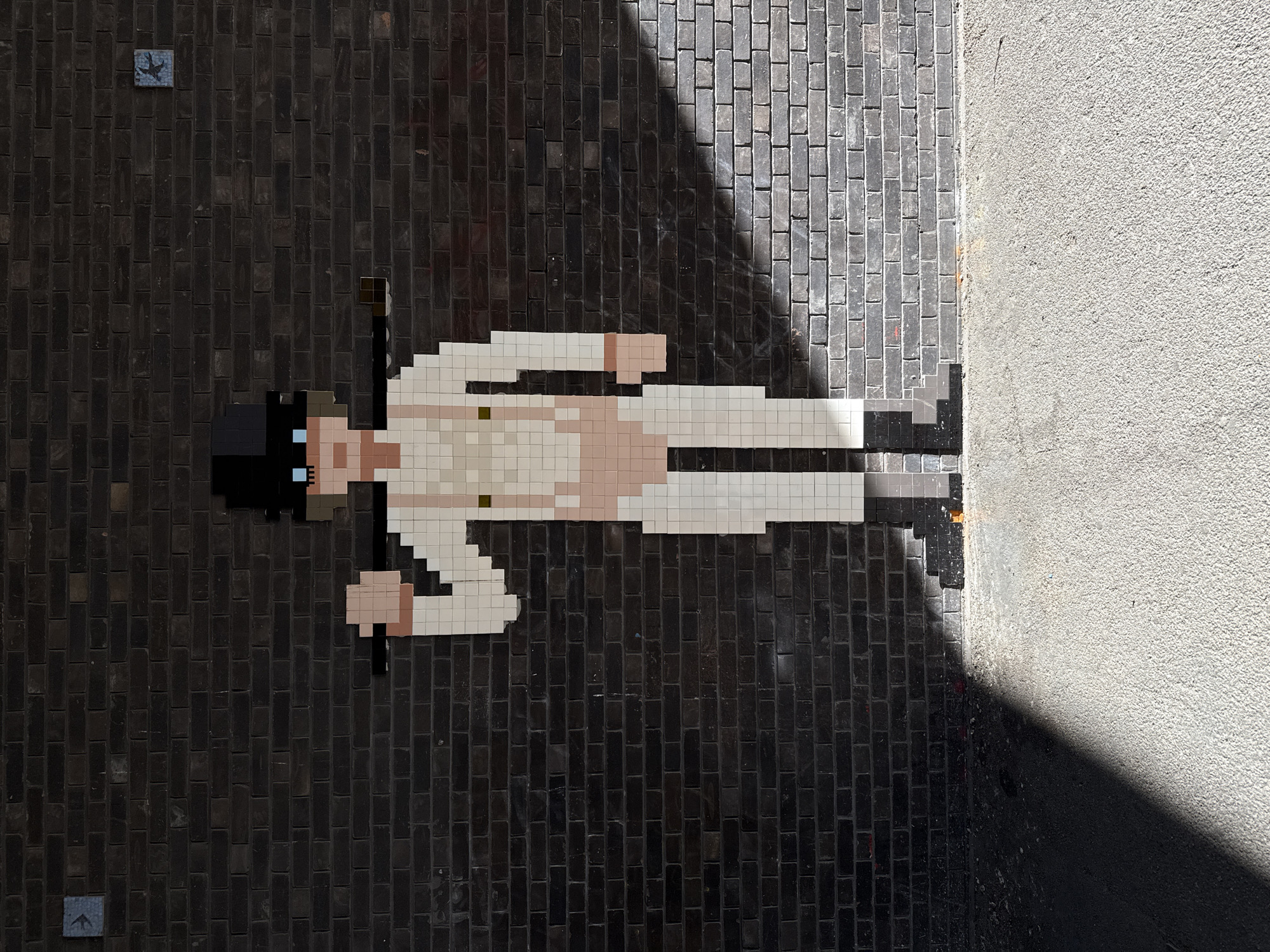A street art mosaic depicting Alex DeLarge from the film "A Clockwork Orange" is affixed to a brick-paved surface. The mosaic figure wears a white outfit, a black bowler hat, and black boots and holds a cane in each hand. The mosaic is made of small, square tiles, giving it a pixelated appearance. To the right of the mosaic, a section of a textured wall is visible, casting a shadow over part of the brick pavement. A small tile with a stylized bird is set in the brick pavement near the upper left corner of the mosaic. This work by Invader is situated in Shoreditch, London, United Kingdom.