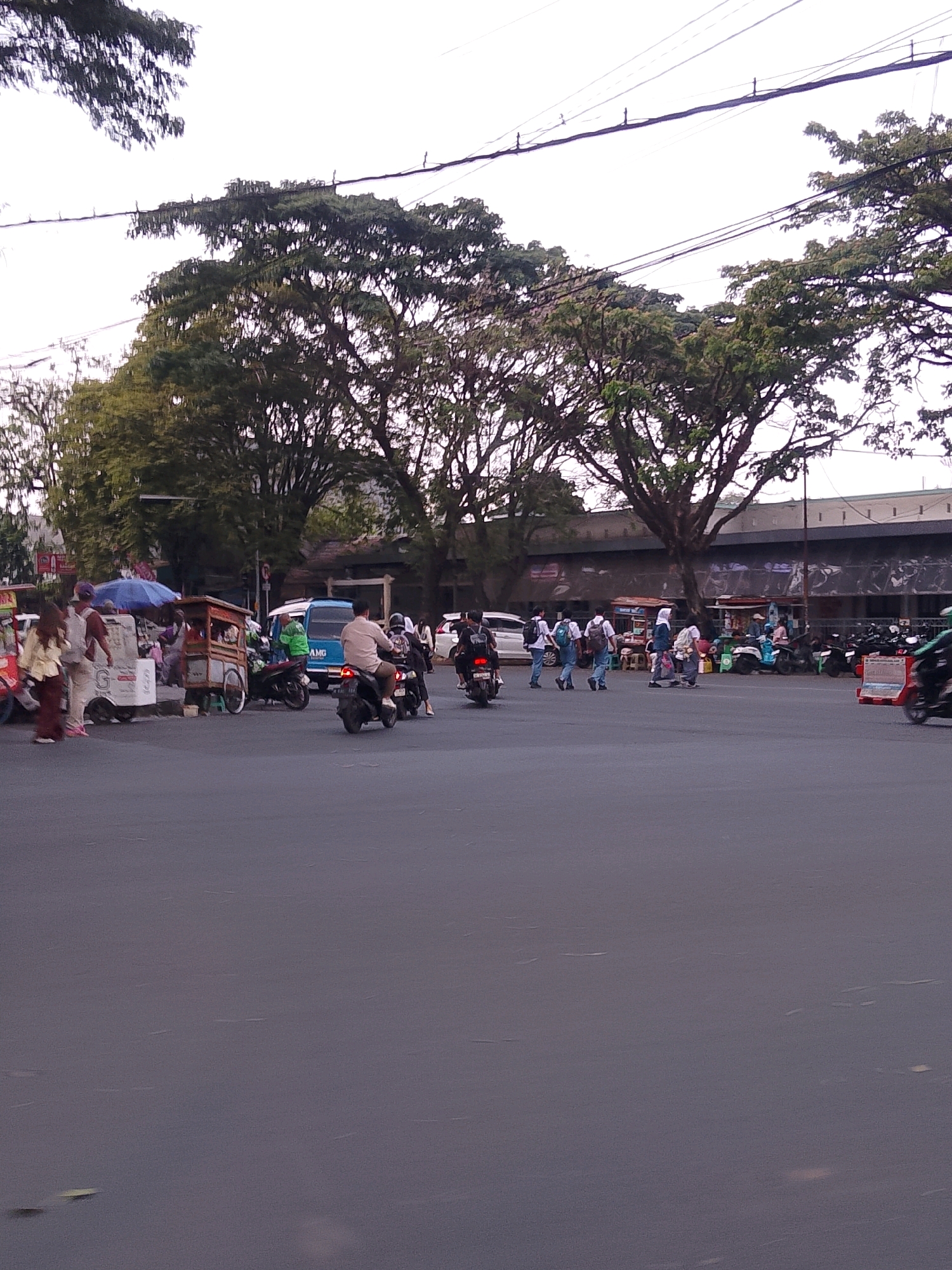 This is an image of a busy street intersection in the city of Malang, Indonesia, likely near a marketplace or commercial area. The foreground is dominated by a grey asphalt road with some reflections. In the middle ground, there is a diverse mix of activity. Several people are walking and standing on the sidewalk, some near food carts, indicated by wooden structures with wheels. One food cart, in particular, is brown with a canopy. A blue van is parked on the side of the road, and several motorcycles are also present, some with riders. One person is riding a motorcycle with a beige shirt, and another is on a black motorcycle. Further back, a long, low building with a dark-colored roof and a white facade stretches across the scene. Large, leafy green trees with prominent branches are visible behind the building and also line the street, with power lines strung between them and poles. The sky is overcast and bright, suggesting it might be late afternoon or early morning. The presence of food carts, a variety of vehicles including motorcycles and a van, and pedestrians suggests a lively urban environment.