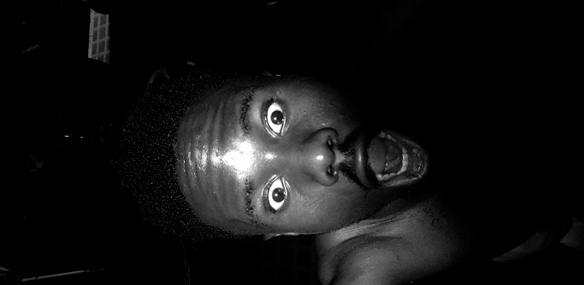 A low-angle, close-up, monochrome photograph depicts a person's face with their mouth open. The person has dark skin, short, dark hair, and appears to be looking directly at the camera. The image is dimly lit, with reflections visible on the person's forehead and face. The environment surrounding the person is dark and indistinct. The location is Narayi, Nigeria.