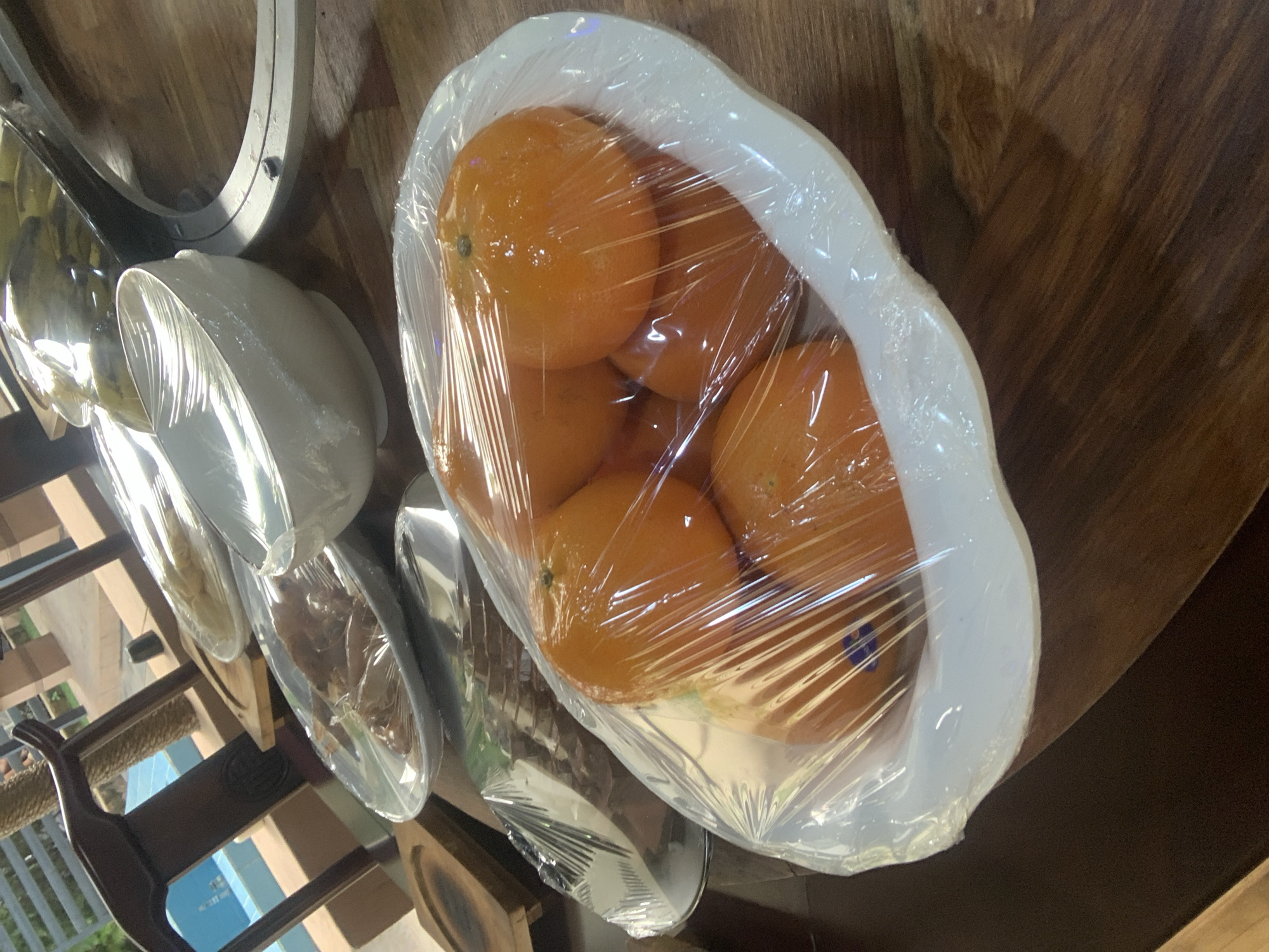 This is a close-up, high-angle shot of a white oval bowl filled with tangerines, covered in clear plastic wrap. The tangerines are a vibrant orange, and their skins appear slightly moist, reflecting the light. A faint purple sticker is visible on one of the tangerines. The bowl sits on a dark wooden table with a visible grain. In the background, out of focus, are other serving dishes covered in plastic wrap, including what appears to be a clear dish with food items and a large white bowl. A portion of a dark wooden chair and a railing are also visible, hinting at an outdoor setting. The scene suggests food preparation or presentation, possibly for an event or a gathering. The presence of other serving dishes and furniture suggests a domestic or hospitality setting within the city of Abuja, Nigeria.