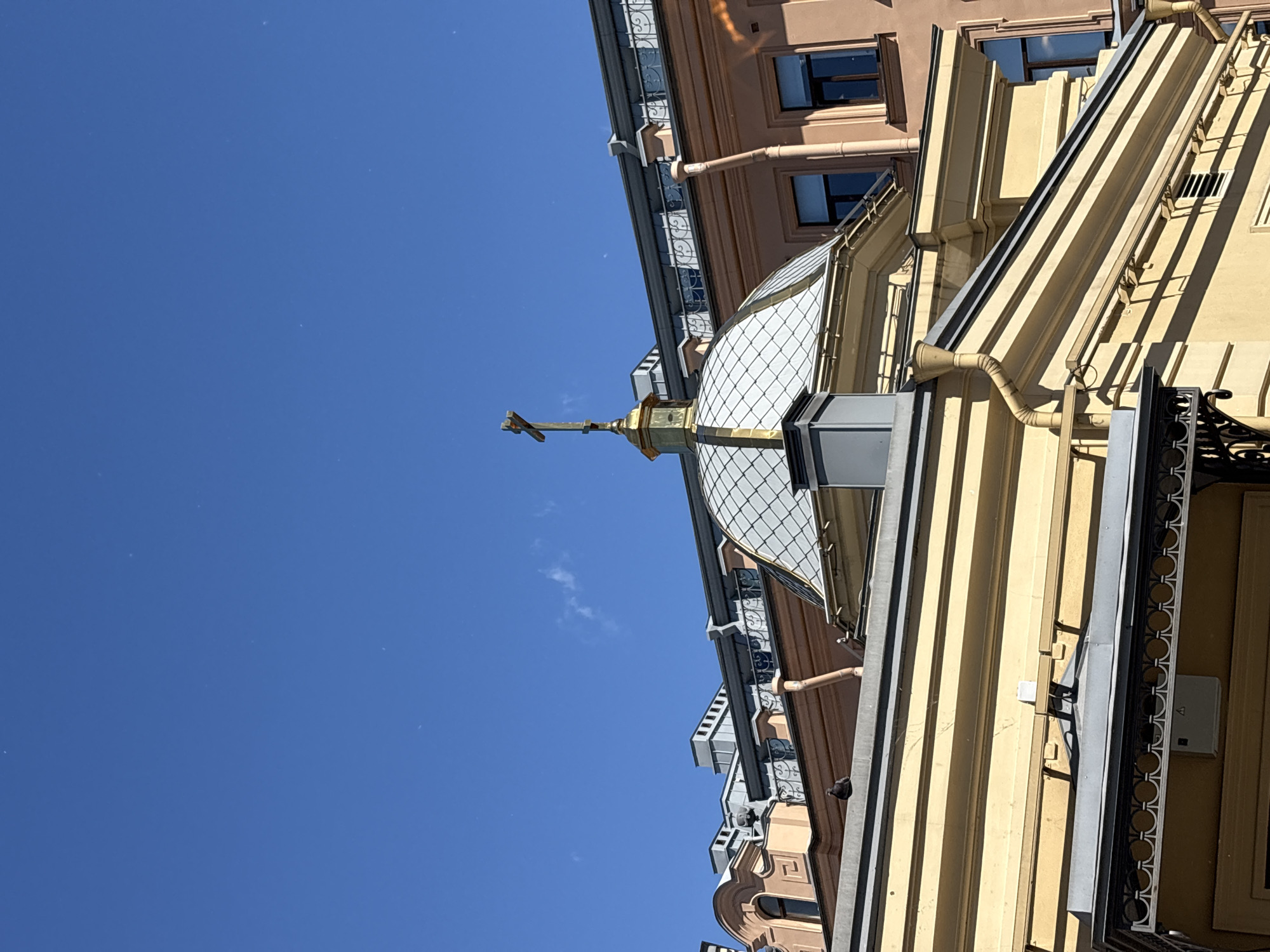 The image shows the roofline of several buildings in Saint Petersburg, Russia, under a clear blue sky. A prominent feature is a domed structure with a cross on top, suggesting it's part of a church or religious building. The dome is topped with a golden cross. Adjacent to the dome are buildings with peach-colored and yellow facades, featuring various architectural details like windows, decorative cornices, and downspouts. A pigeon is perched on the ledge of one of the buildings. The overall scene depicts a section of the urban landscape characteristic of Saint Petersburg's historical architecture.