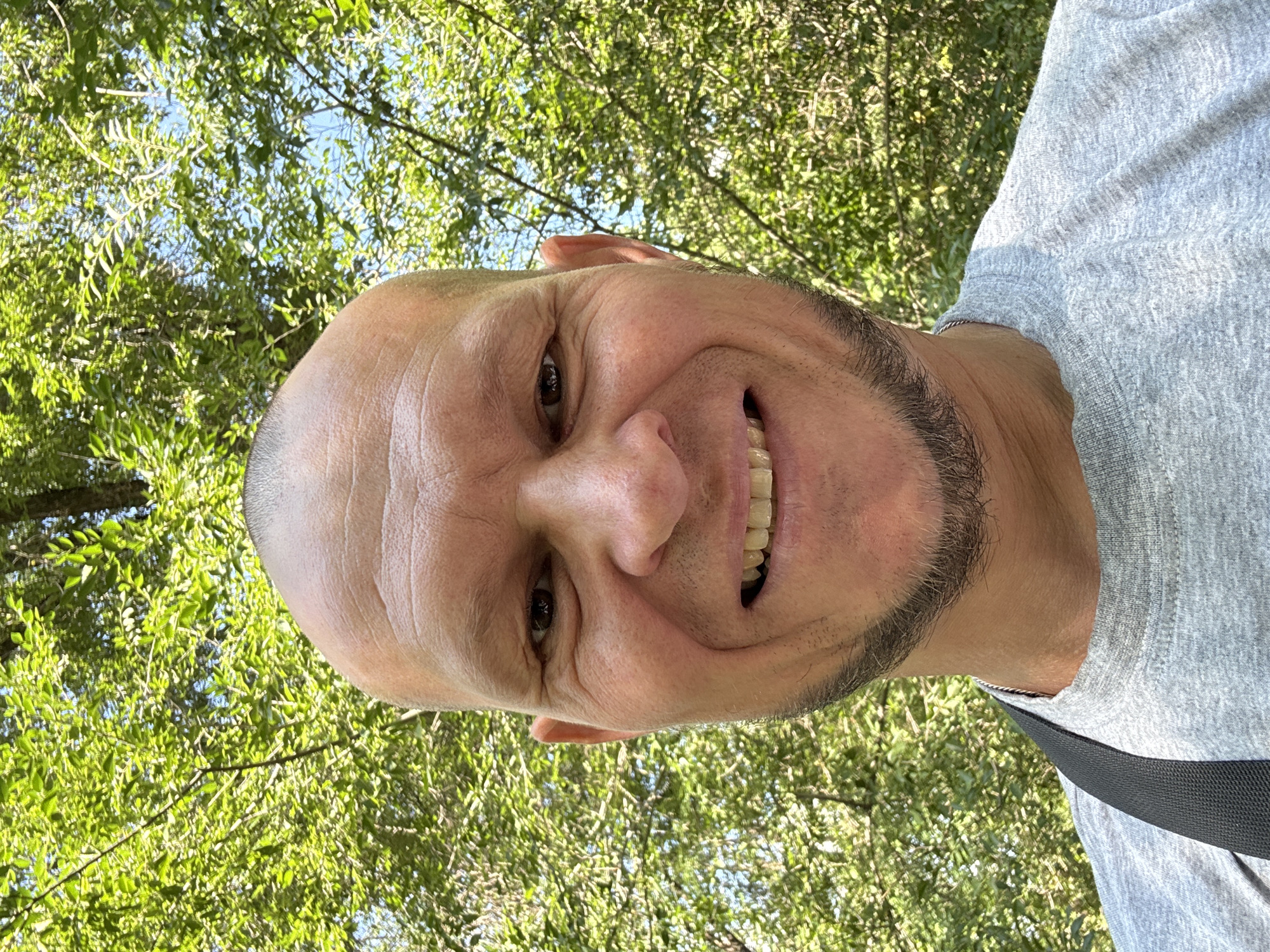 A man with a shaved head and a goatee is shown in a close-up selfie taken outdoors. He is wearing a grey t-shirt and has a black strap across his shoulder. The background is filled with green foliage and dappled sunlight, suggesting a park or wooded area. The man is smiling, with his teeth visible. The image quality is clear and sharp. There is no visible information indicating the location in Oskemen, Kazakhstan.