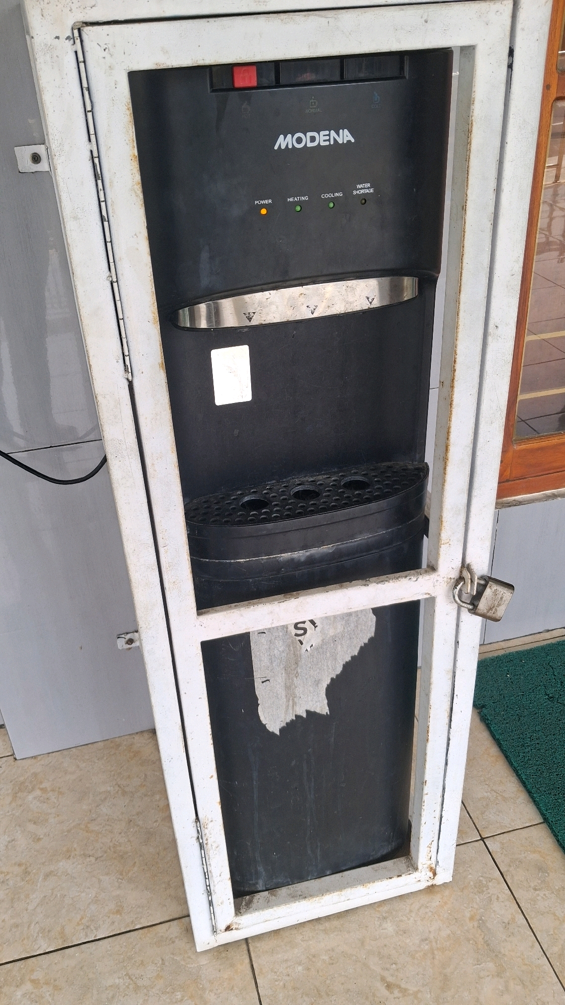 This is a medium shot, eye-level, outdoor, full-body shot of a black water dispenser for sale at an outdoor market in Giriwarno, Indonesia. The water dispenser is a standard upright model with a black plastic exterior and silver trim around the dispensing area. It is housed within a white metal cage with a hinged door secured by a padlock. The dispenser has indicator lights for "POWER," "HEATING," "COOLING," and "WATER SHORTAGE." The "POWER" light is orange, and the "HEATING" and "COOLING" lights are green. The "WATER SHORTAGE" light is off. The dispenser has a removable black drip tray with circular openings. The base of the dispenser is a large black cylinder, possibly a water tank, with torn paper attached to its surface. The cage is attached to a gray wall on the left and a wooden-framed window on the right. The ground is tiled with light brown square tiles, and a green mat is visible to the right. The overall scene suggests a commercial setting where a used appliance is being offered for sale.