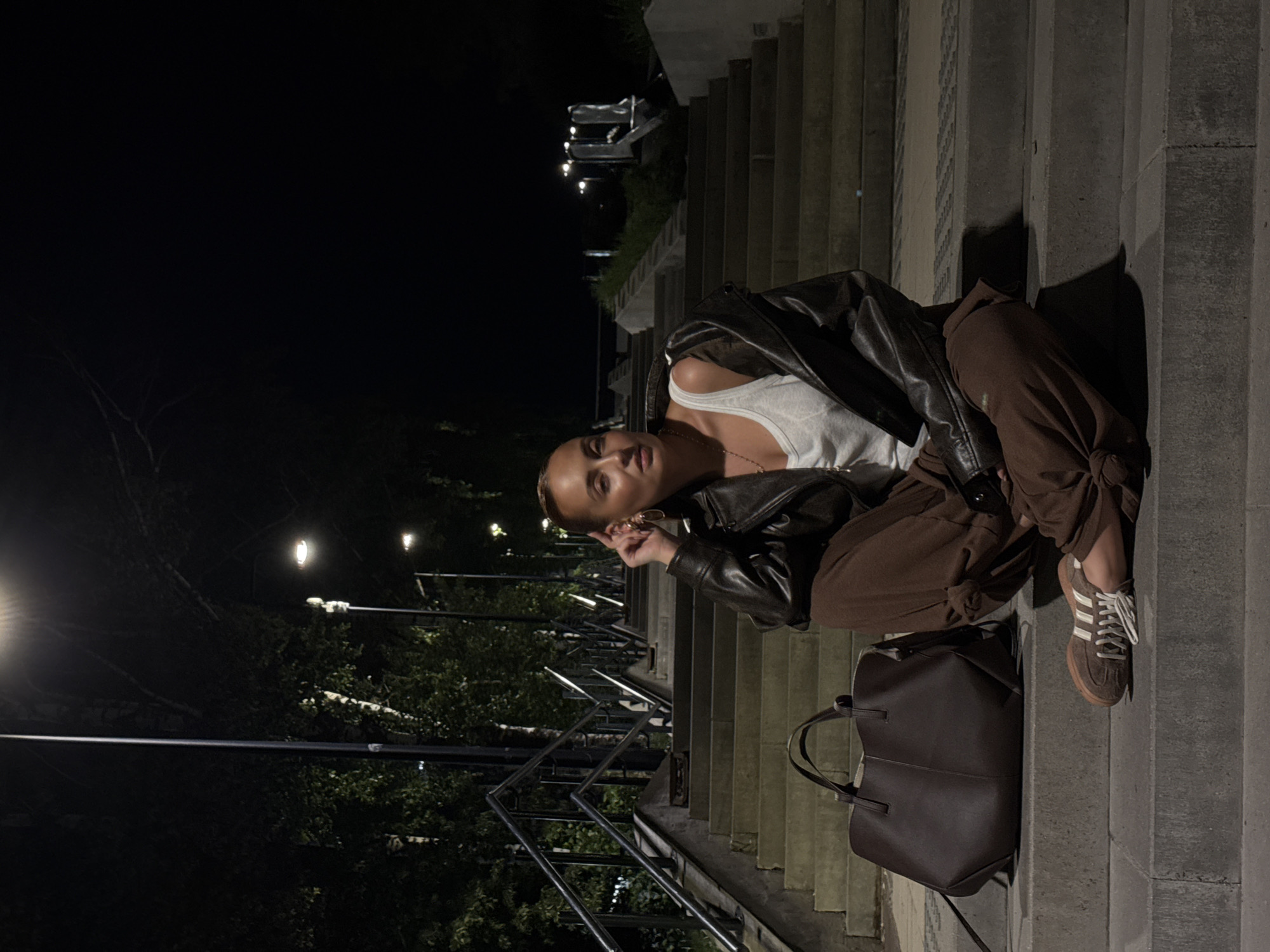 A woman is sitting on concrete steps outside at night, potentially in Izhevsk, Russia, based on visual cues of urban landscape and architectural style. She is wearing a white tank top, brown pants, a dark leather jacket, and brown sneakers with white stripes. A dark brown tote bag is on the step next to her. The steps lead up to an area illuminated by streetlights, with trees visible in the background. Metal railings run along the side of the steps. The lighting is dim, with streetlights providing the main source of illumination.