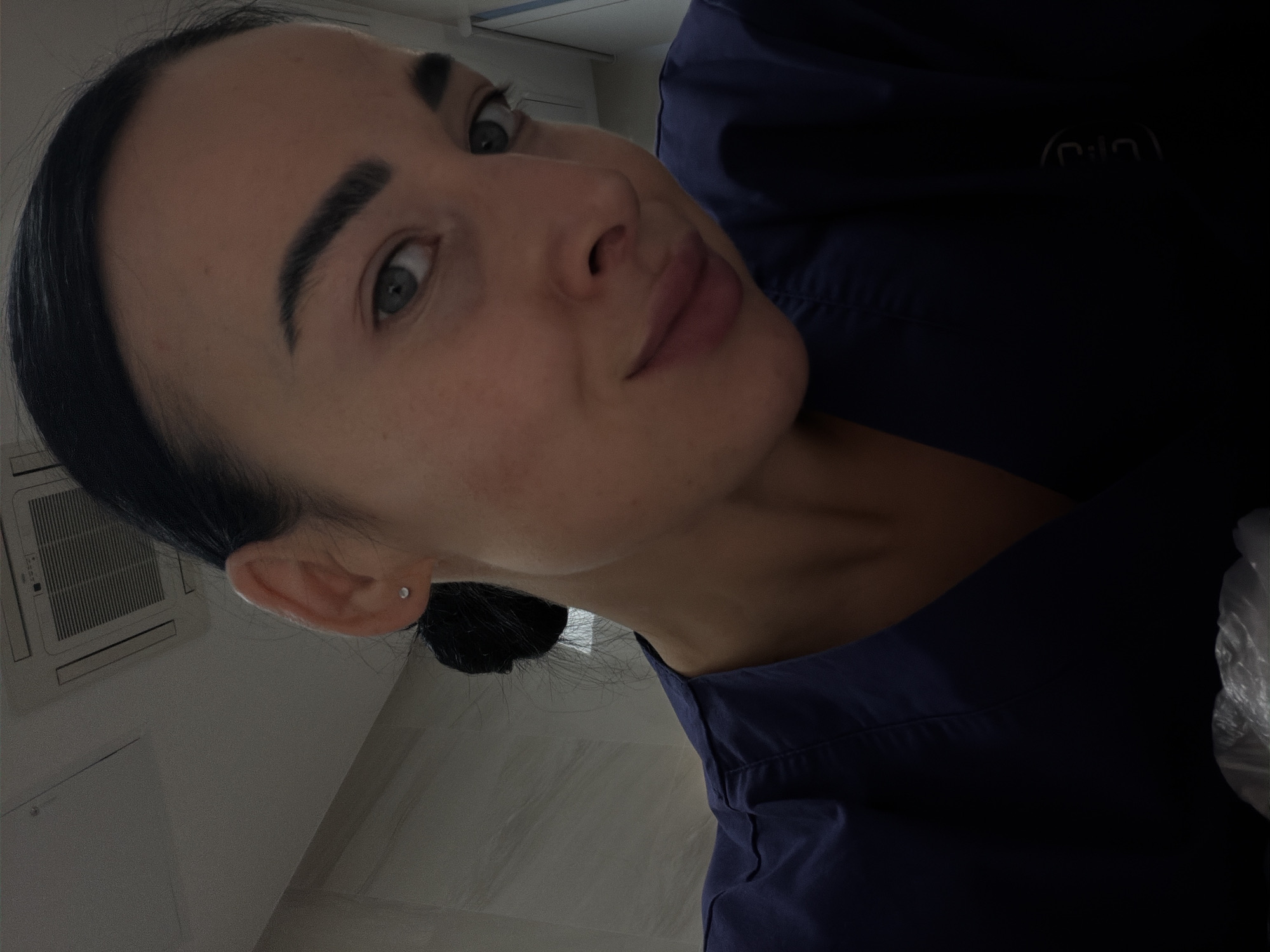 A medium close-up shot shows a woman wearing a dark blue or black uniform, possibly scrubs, in a brightly lit, sterile-looking environment. The lighting seems to come from overhead, illuminating her face, which is turned slightly upward and to the left. She has dark hair pulled back into a bun, prominent dark eyebrows, and light-colored eyes. Her expression is subtle, almost a slight smile. A small, silver earring is visible in her left ear. In the background, a white air conditioning unit is mounted on the wall. The surroundings suggest a medical setting, such as a clinic or hospital. There is no information to determine if the location is in Saint Petersburg, Russia.