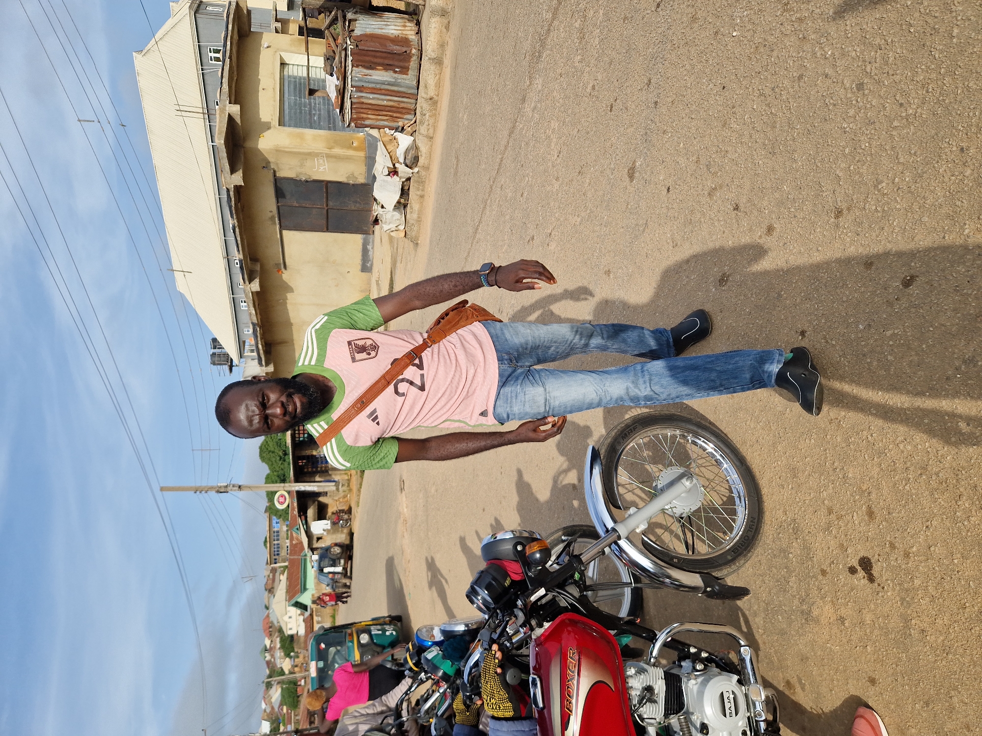 A medium-shot, full-body, vertical photograph taken outdoors in the city of Kabong, Nigeria, captures a man walking towards the camera. The man has dark skin and a beard, and he is wearing a pink t-shirt with green stripes on the sleeves and a "24" printed in the center, blue denim jeans, and black shoes. A brown satchel is slung across his chest.
To the man's left, a red and silver motorcycle with the brand name "BOXER" is parked. Its chrome fender and wheel are visible. Further to the left, a yellow and green auto-rickshaw is also parked. In the background, several buildings with corrugated metal and tiled roofs are visible, along with power lines crisscrossing the sky. The ground is paved with asphalt, and shadows of people and objects are cast across it, indicating a sunny day. The scene depicts a street in a residential or commercial area of Kabong, Nigeria.
