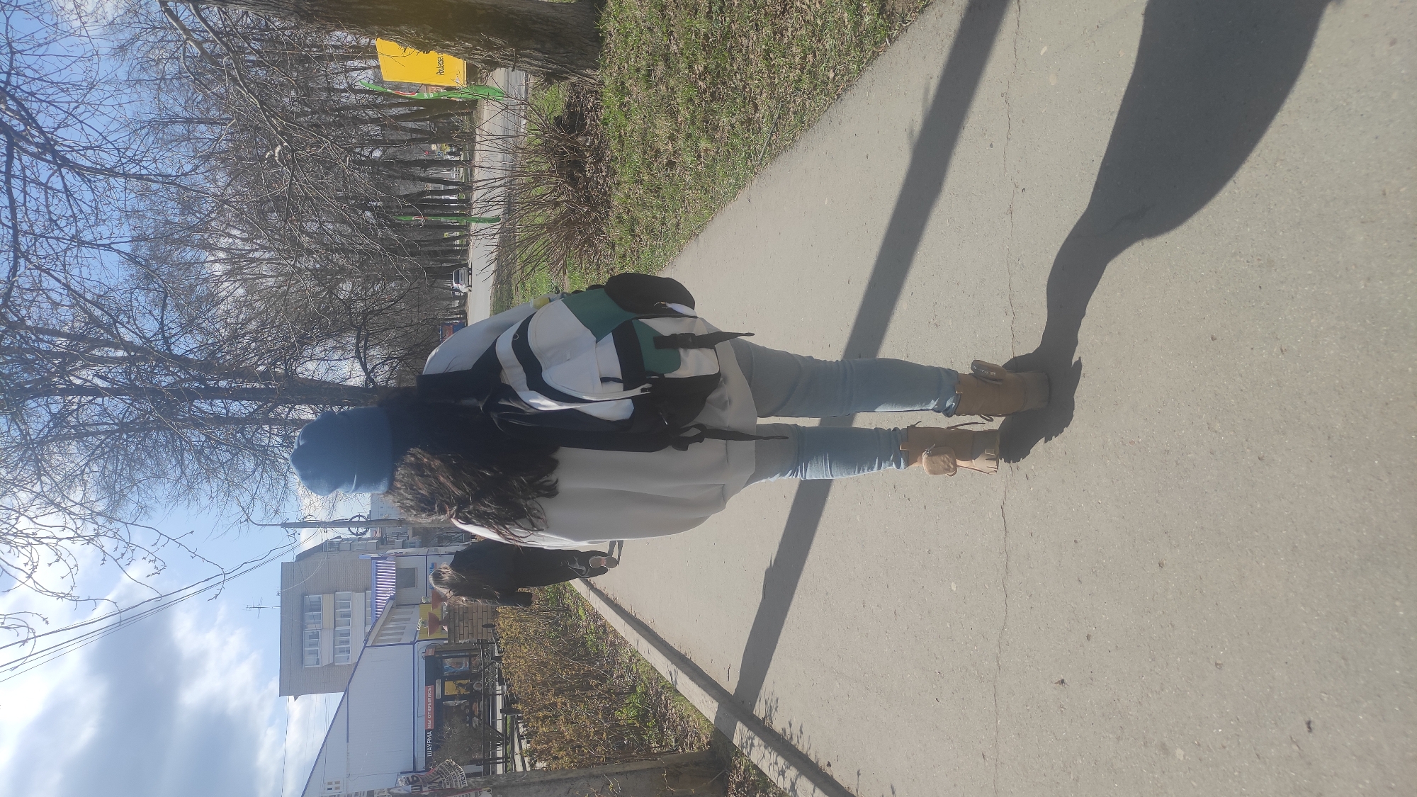 The image captures an outdoor scene in Chaikovsky, Russia, showing a person walking away from the camera on a paved sidewalk. The person is wearing light-wash blue jeans, brown lace-up boots, a white or light-colored hoodie, a backpack with a black base, white main flap, and green accents, and a blue beanie. Long dark hair is visible beneath the beanie. A dark, fluffy, dog-like animal with a fluffy tail is partially visible, walking close beside or slightly behind the person.
The sun is low in the sky, casting long, dark shadows of the person and other objects onto the pavement, suggesting either early morning or late afternoon. The sky is bright blue with scattered white clouds. To the left of the path, an area with numerous bare trees stretches into the distance, indicating late winter or early spring. Beyond the trees, a street with a distant car and various buildings are visible. One prominent building in the mid-ground appears to be an apartment block or commercial structure.
Visible Russian text on a distant yellow sign among the trees likely reads "Продается" (For sale). Blurry Russian text on a sign attached to a building in the background appears to indicate "ШАУРМА ВЫПЕЧКА" (SHAURMA BAKERY), suggesting food establishments. Power lines traverse the sky in the mid-ground. The ground adjacent to the path is covered with sparse grass and dry vegetation.