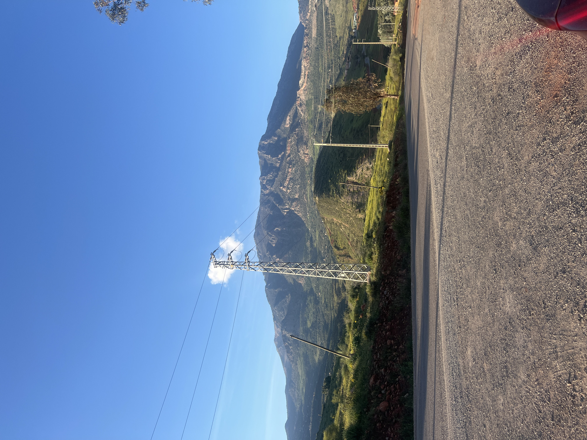 The image shows a rural roadside scene in Ouaouizert, Morocco. A paved road runs vertically on the right side of the image. A mountain rises in the center, covered with vegetation. A power transmission tower stands on the left, with power lines extending across the sky. A clear blue sky is visible in the upper-left corner. The ground is a mix of dirt, rocks, and green vegetation. Part of a vehicle is visible in the upper right.