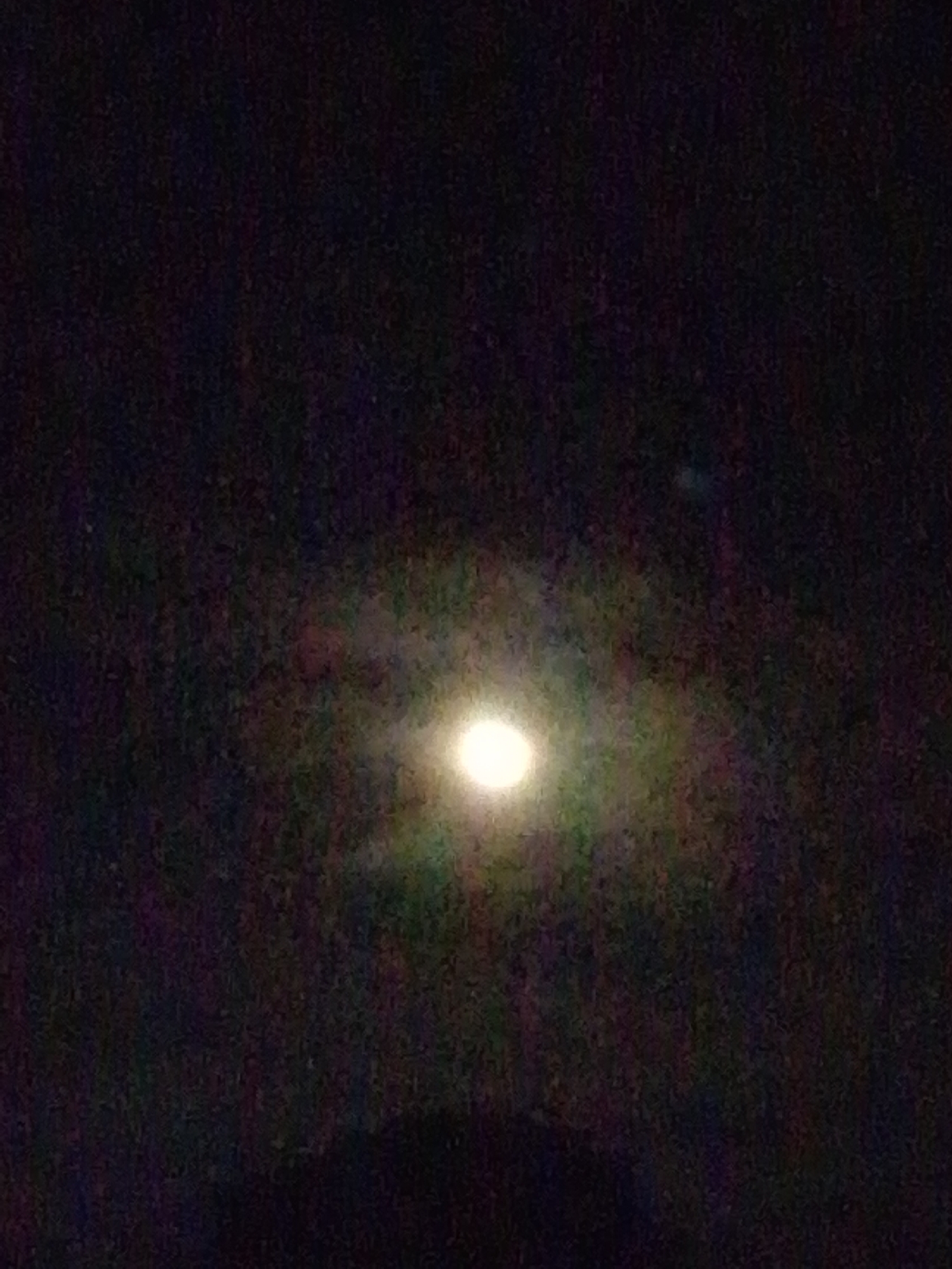 The image is a dark, grainy photograph with a bright, circular object in the center, likely the moon. The background is black with some faint, colorful haziness around the central object, suggesting atmospheric conditions or lens flare. The image appears to have been taken at night. There is no discernible location in the city of Serti, Nigeria visible.