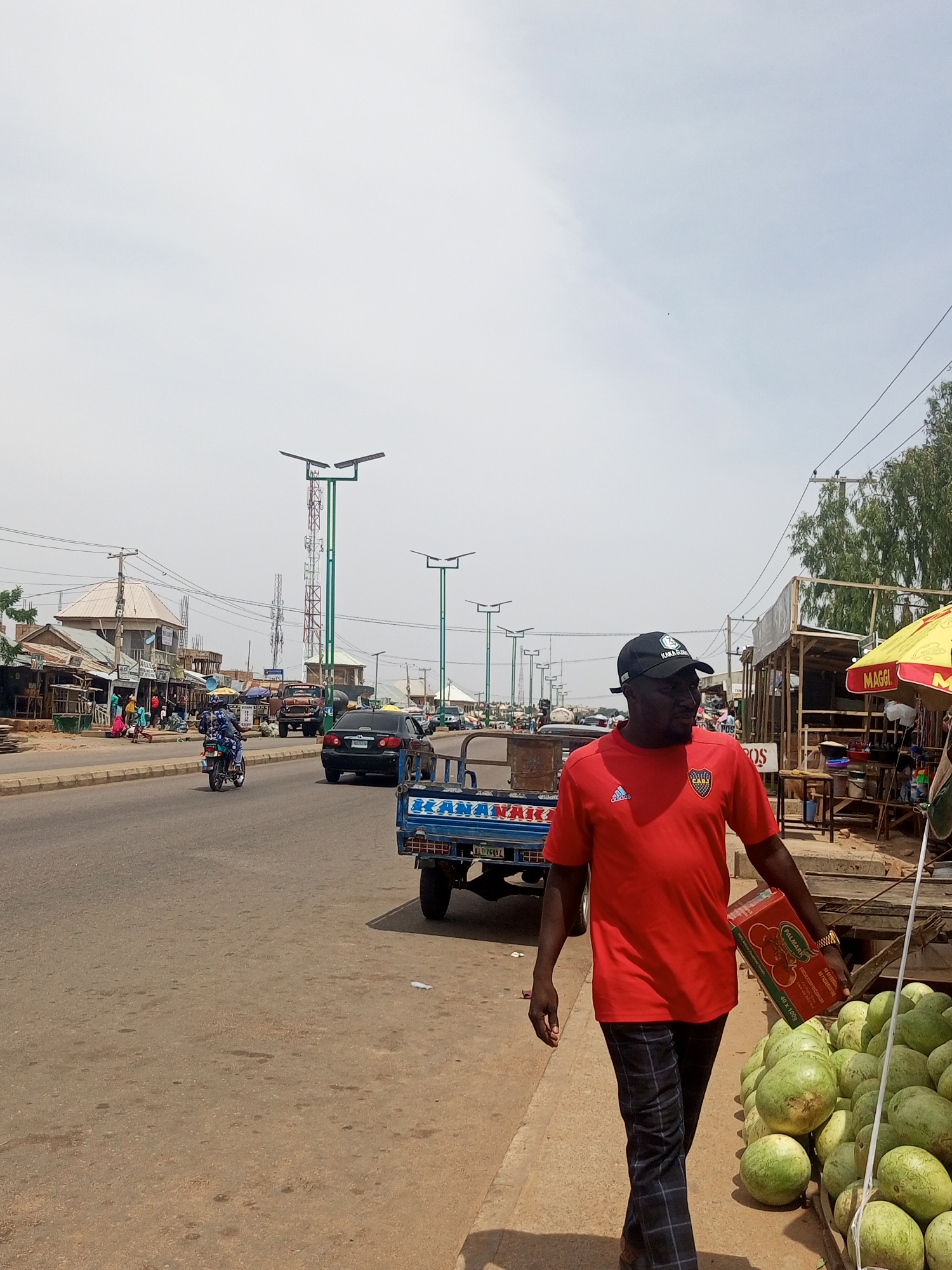 The image depicts a street scene in Tempure, Nigeria, with a focus on a man walking along the roadside. The man, wearing a red t-shirt and checkered trousers, carries a box of what appears to be tomatoes. He is walking past a stall selling watermelons, a common sight in Nigerian markets. The street is paved and has a sidewalk. In the background, there are various shops and market stalls, and a truck is visible on the road. Several streetlights are also present, indicating a developed urban area. The sky is bright and cloudy, suggesting a warm day. The scene captures the hustle and bustle of a Nigerian street market.