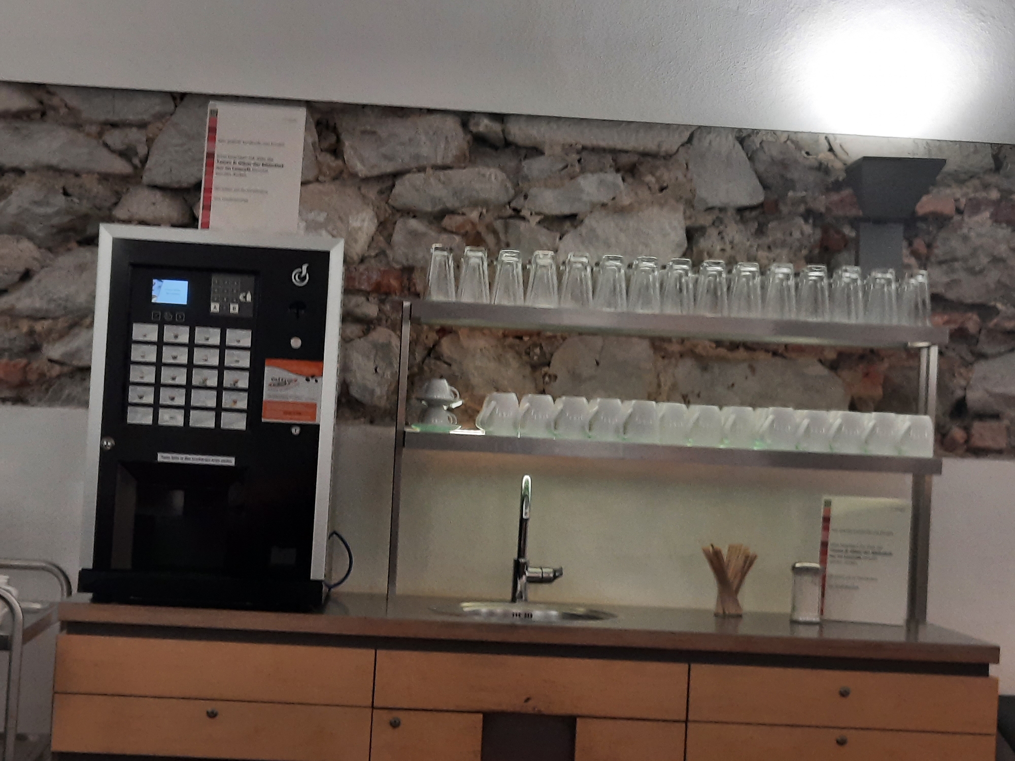The image depicts a coffee machine and a sink area set against a stone wall. The coffee machine is a white and black appliance with a digital display and numerous buttons. Above the coffee machine, a sign with red and white text is attached to the wall. To the right of the coffee machine, a shelf holds multiple rows of clear glasses. Below this shelf, a stainless steel sink with a modern faucet is visible. On the countertop next to the sink, there are wooden stirrers in a holder, a small shaker, and another sign with text. The scene appears to be in a public or commercial establishment. The presence of a coffee machine and sink suggests a self-service area for beverages. The overall impression is one of a functional and simple setup, likely found in a café, break room, or similar location. Based on the provided information, this scene is located in Rosenheim, Germany.