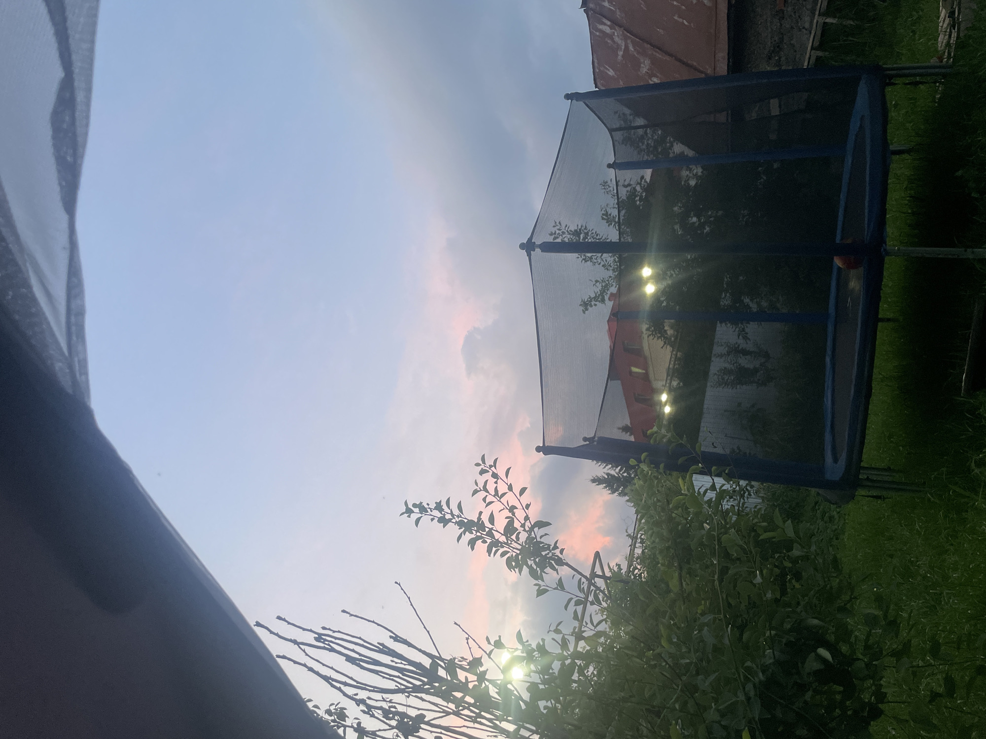 The image depicts an outdoor scene at dusk, likely in Laishevo, Russia, as suggested by the context. In the foreground, lush green grass covers the ground, with a trampoline positioned prominently in the center. The trampoline is blue with a black safety net enclosure and has visible metal legs supporting it. A small orange ball rests on the trampoline's mat. To the left of the trampoline, a dense green bush with leaves and branches is visible, and beyond that, a tree with bare branches extends upwards, catching the last light of the sun. The sun itself is partially visible behind the foliage on the left, emitting a strong glare. In the background, a red-roofed building with illuminated windows is situated behind the trampoline and trees. The sky above is a mix of pale blue and scattered clouds, with streaks of pink and orange suggesting the fading light of sunset. The overall atmosphere is tranquil and somewhat moody due to the twilight conditions.
